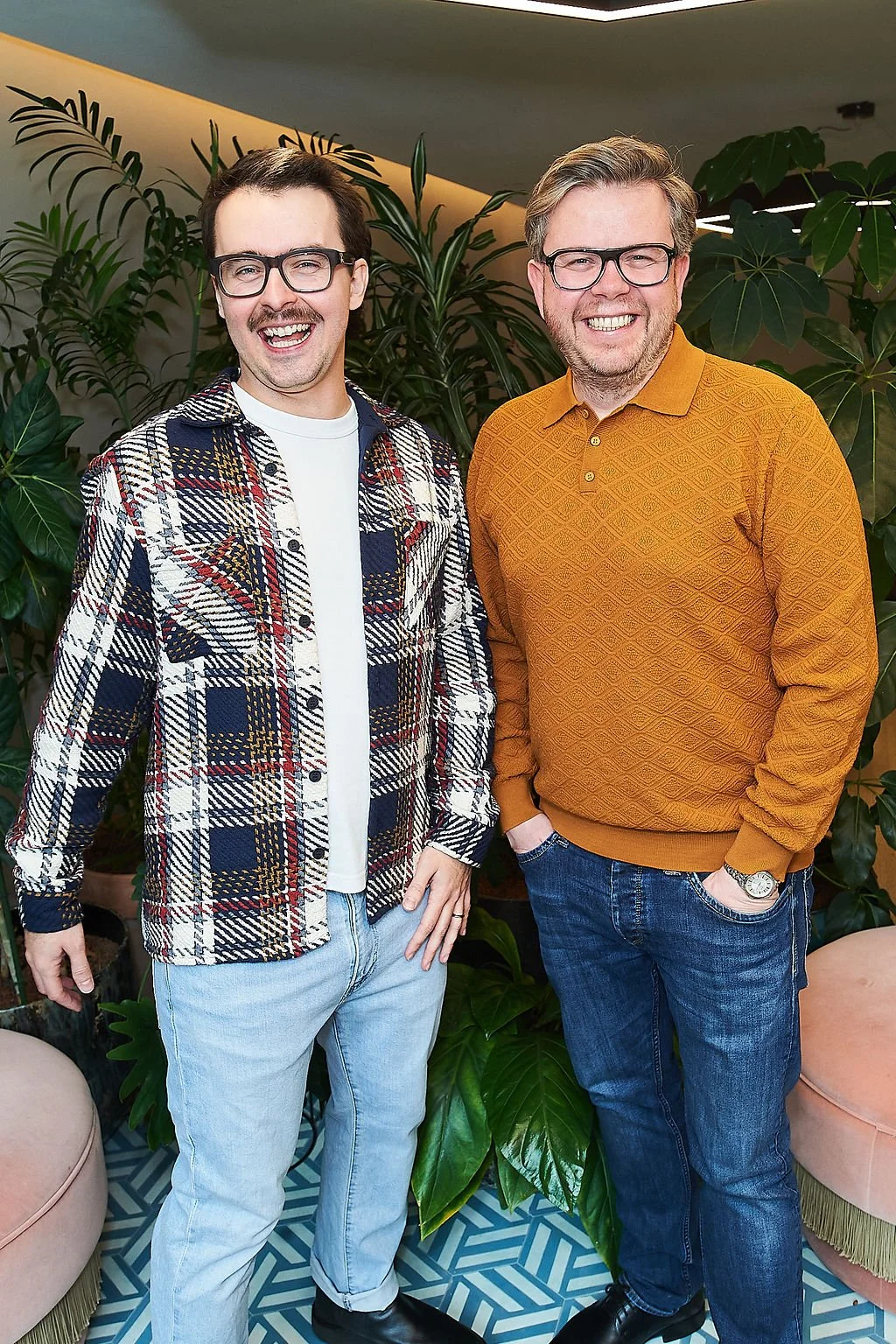 Two men standing together smiling, wearing glasses and casual clothing, in an indoor setting with green leafy plants in the background.