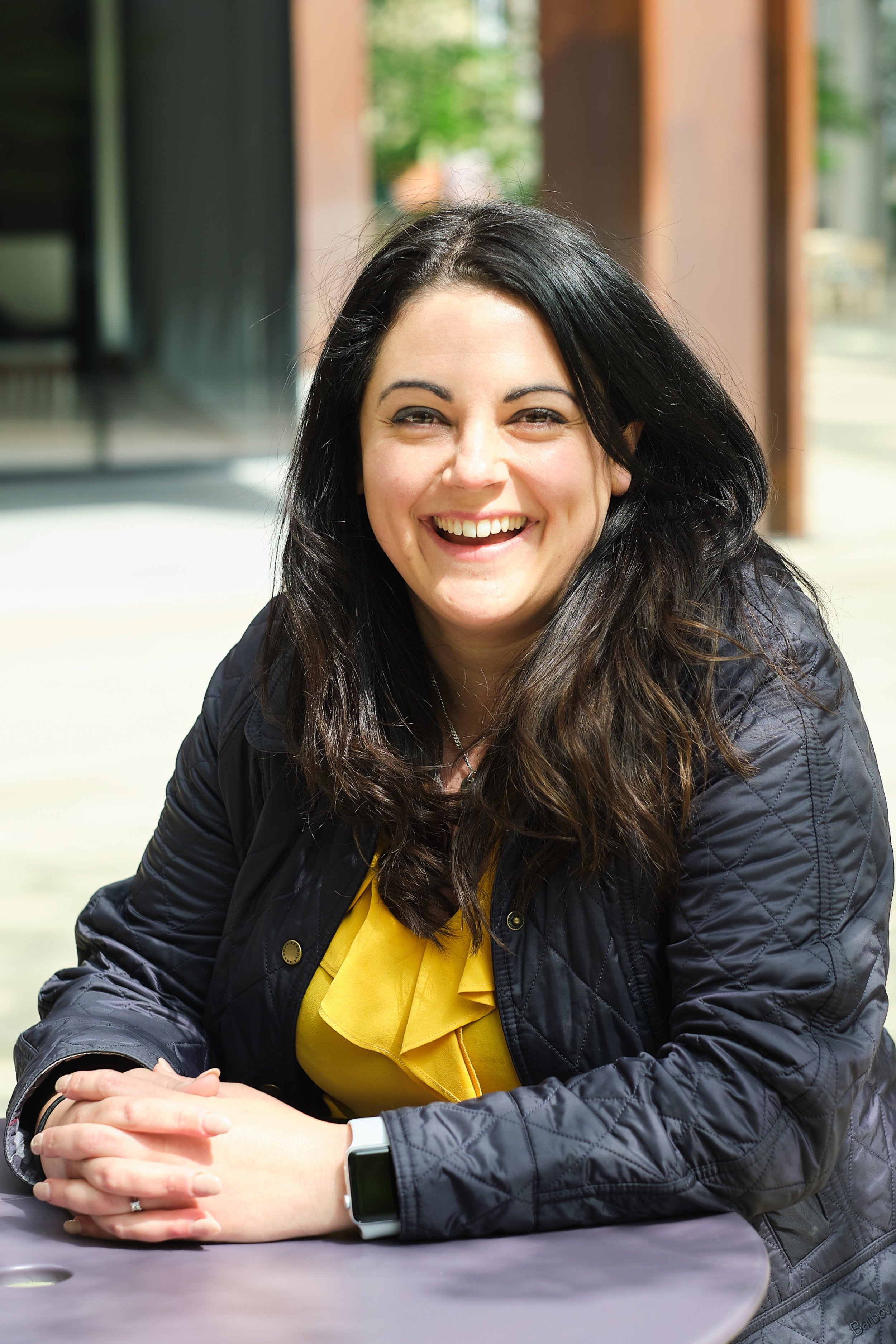 A woman with long dark hair, smiling, sitting at a table outdoors, wearing a black quilted jacket and a yellow top.