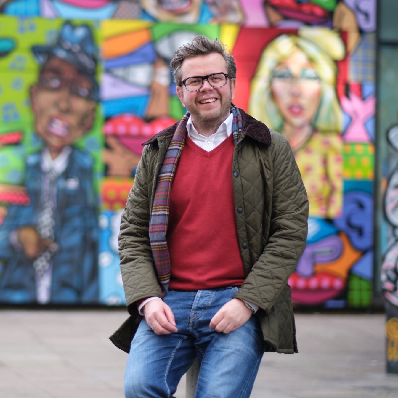 A man with glasses and a beard, smiling, posing in front of colorful graffiti art featuring animated characters.