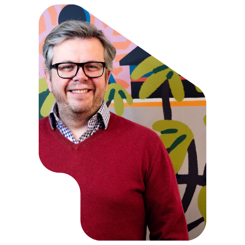 A smiling man with gray hair, glasses, wearing a red sweater over a checkered shirt, standing in front of a colorful mural with stylized palm trees and abstract shapes.