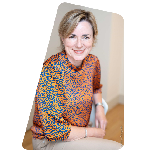 Portrait of a middle-aged woman with light brown hair, smiling, wearing an orange and blue animal print blouse, sitting indoors.