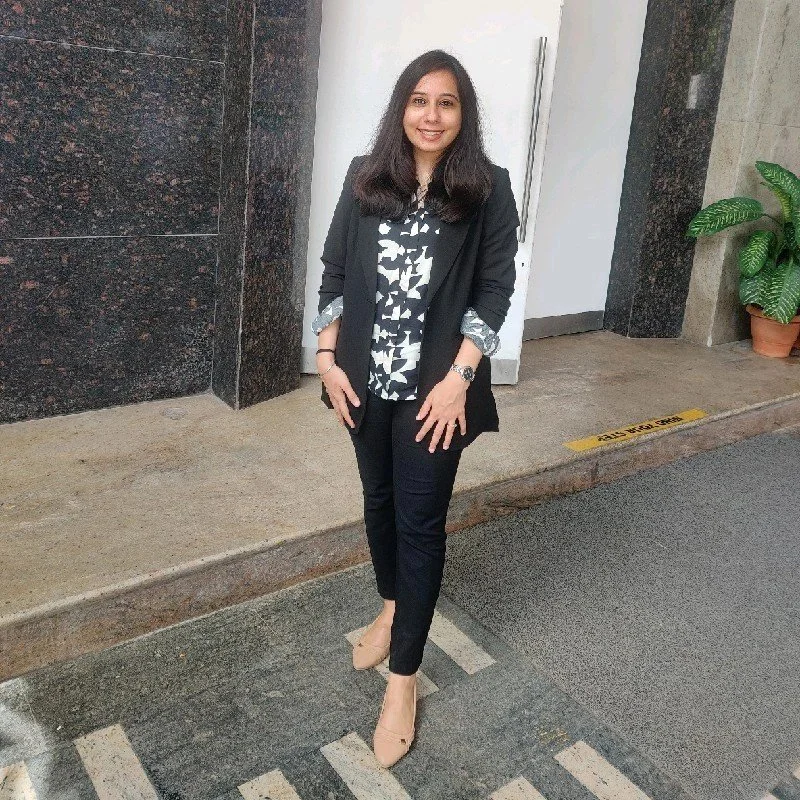 A woman with long dark hair, wearing a black blazer, patterned blouse, black pants, and nude heels, standing on a city street sidewalk with a building and potted plant in the background.
