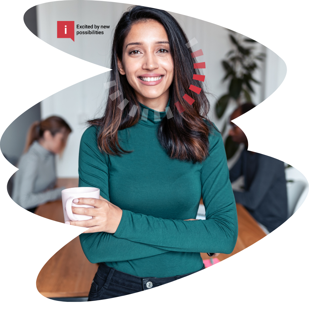 A young woman smiles and holds a white mug in a modern office setting. She wears a teal long-sleeve top and has shoulder-length dark hair. In the background, two colleagues are working at a table, with a potted plant and office furnishings visible.