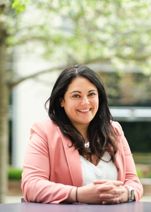 Smiling woman with dark hair wearing a pink blazer and white top, outdoors with trees in the background.