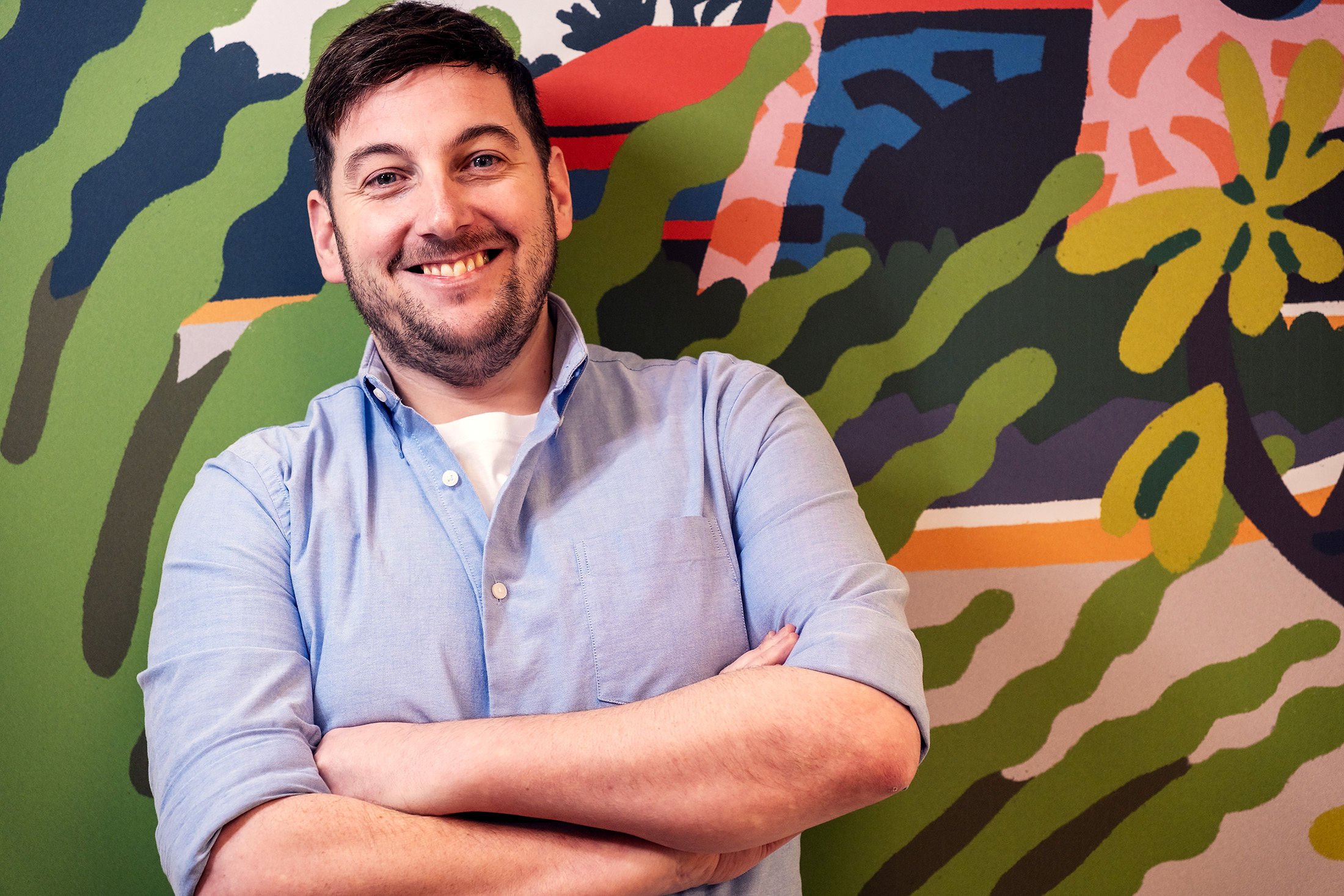 A smiling man with dark hair and a beard, standing with arms crossed in front of a colorful abstract mural featuring shades of green, orange, pink, and blue.