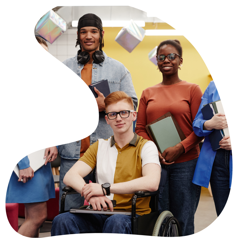 Group of four diverse young adults, two women and two men, smiling in a modern indoor setting. One young man is seated in a wheelchair, holding a laptop, and the others are standing, holding books and notebooks, with a colorful background.