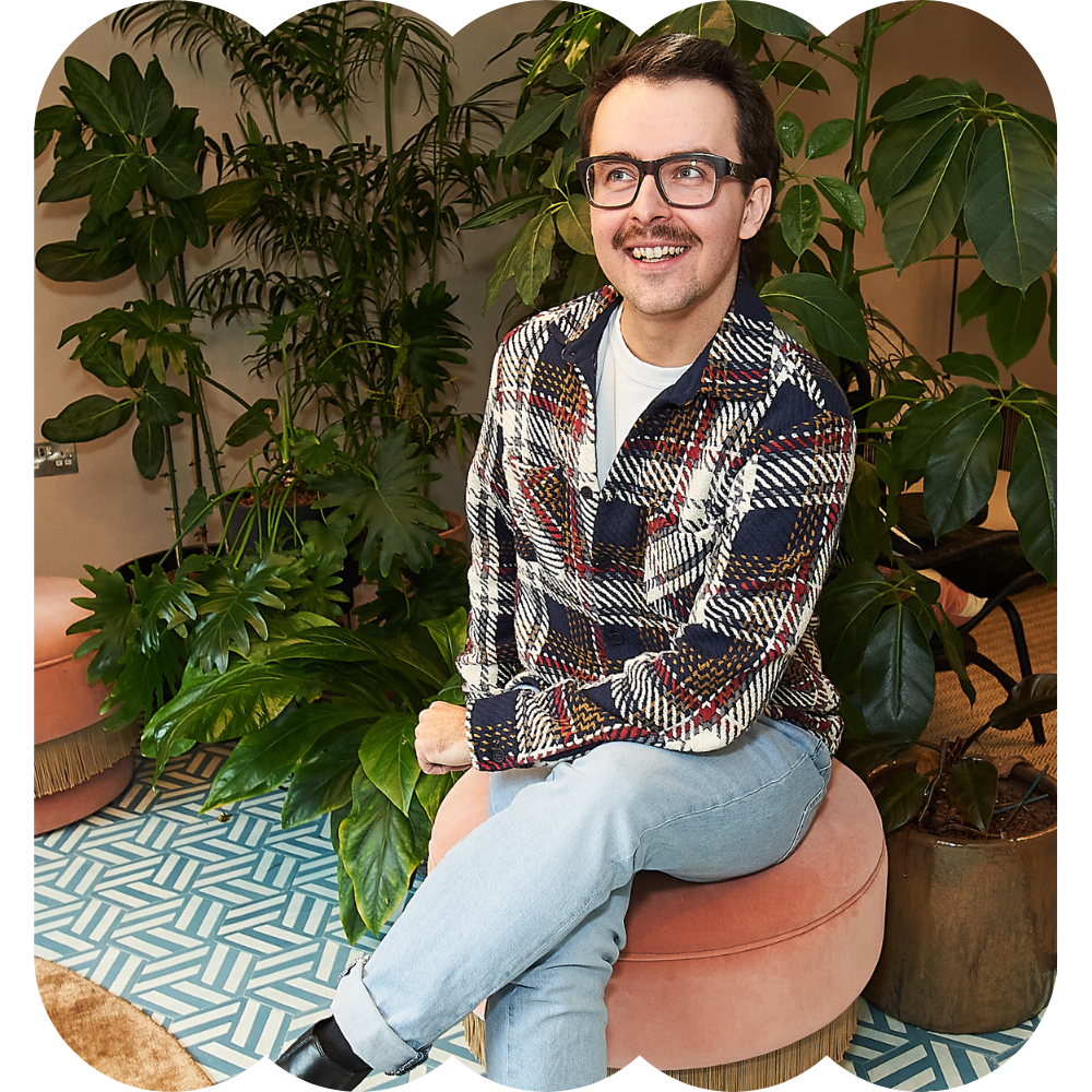 A young man with glasses and facial hair, smiling and sitting on a round pink ottoman, surrounded by large green indoor plants in a cozy, stylish interior.
