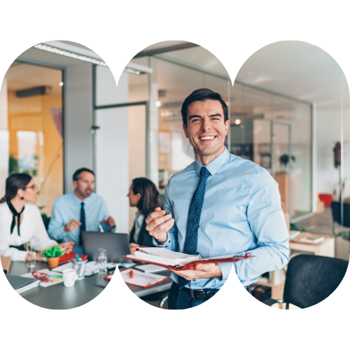 A smiling man in business attire holding a red folder and pen in a modern office conference room, with colleagues sitting at a table engaged in a discussion in the background.