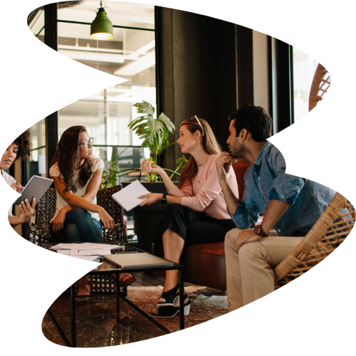 Group of five young adults having a discussion in a modern café or lounge, seated on couches and chairs around a coffee table, with large windows and greenery in the background.