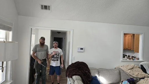 Two men standing in a living room near an open doorway, with a sectional sofa and a cozy blanket on the couch.