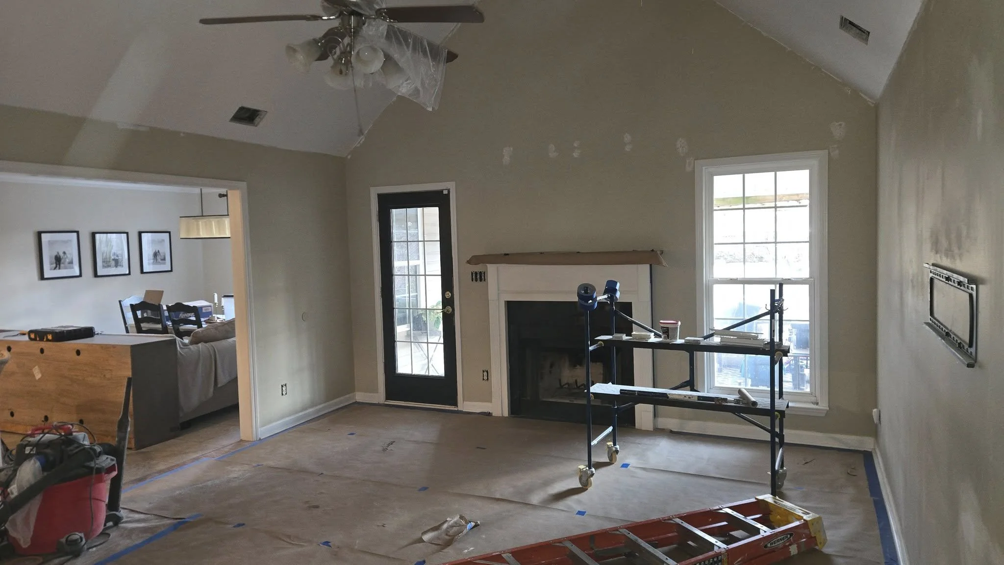 Unfinished living room with a ceiling fan, a fireplace, and large windows. The floor is covered with protective paper, and construction tools and scaffolding are present.