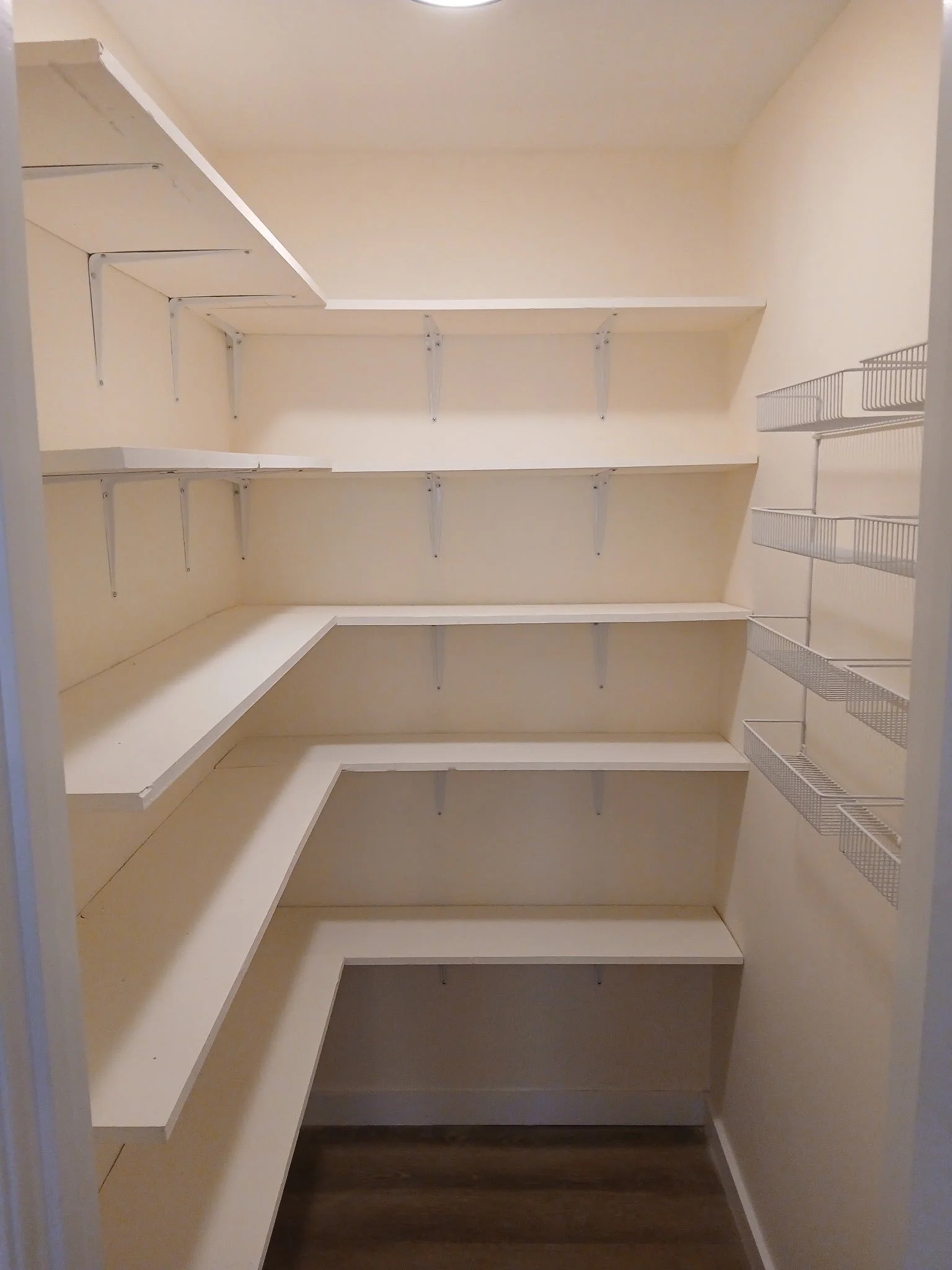 Empty walk-in closet with matching cream walls and shelving units and wire baskets on the right wall.