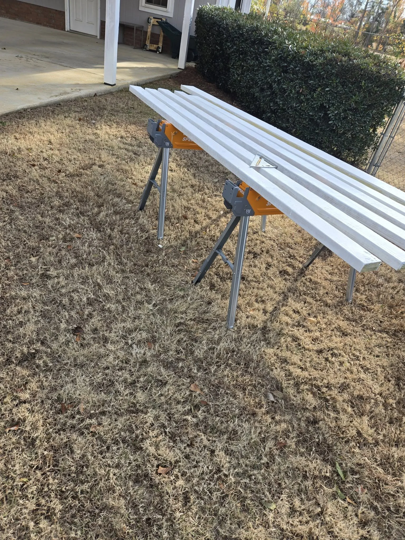 A freshly painted white wooden board is placed on a pair of orange and gray sawhorses on a lawn with brownish grass. In the background, there is a house porch with a green trash bin and some other objects behind a bush and a chain-link fence.