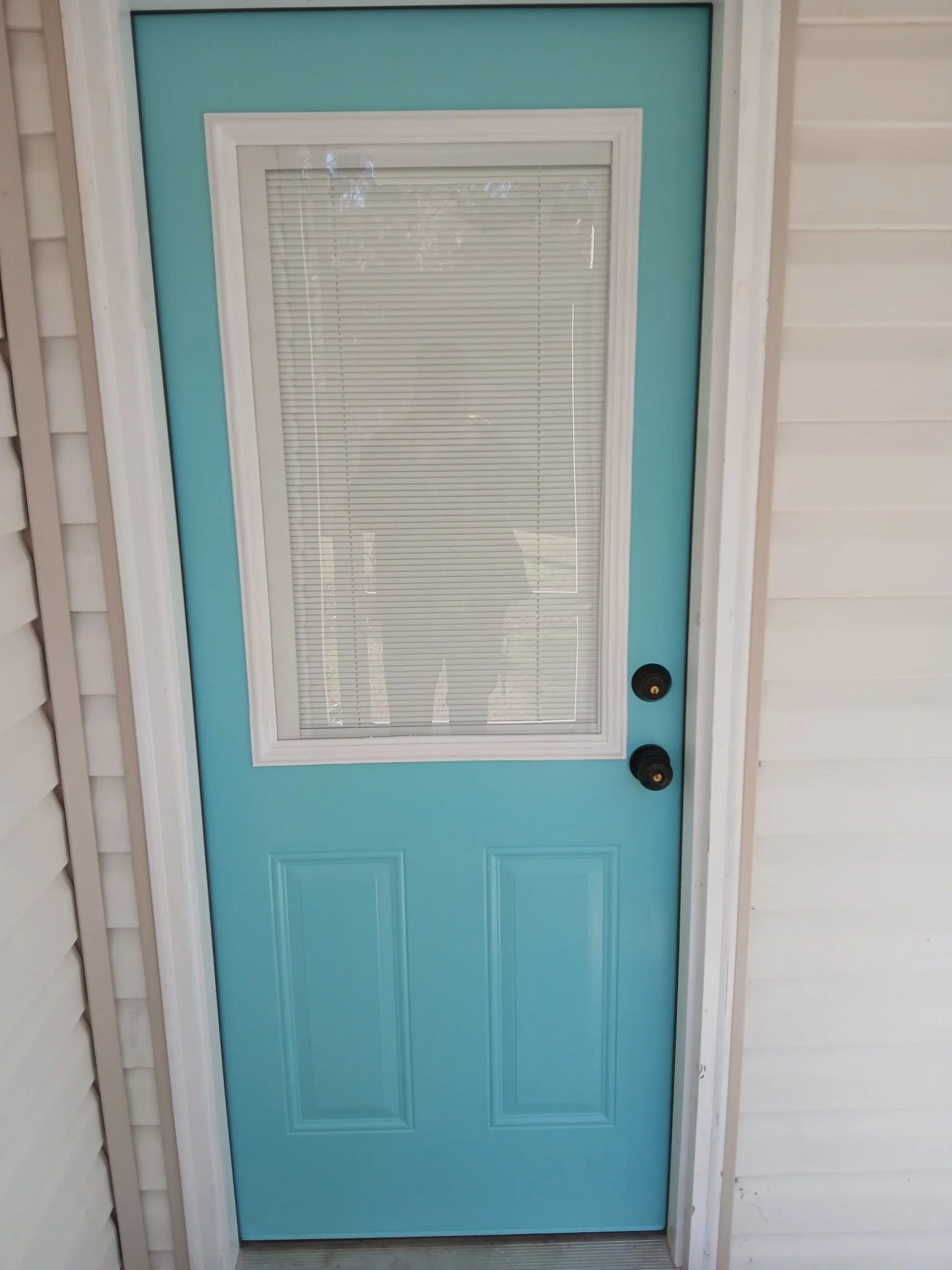 Blue exterior door with a window and blinds, white trim, black doorknobs, white siding on the house.