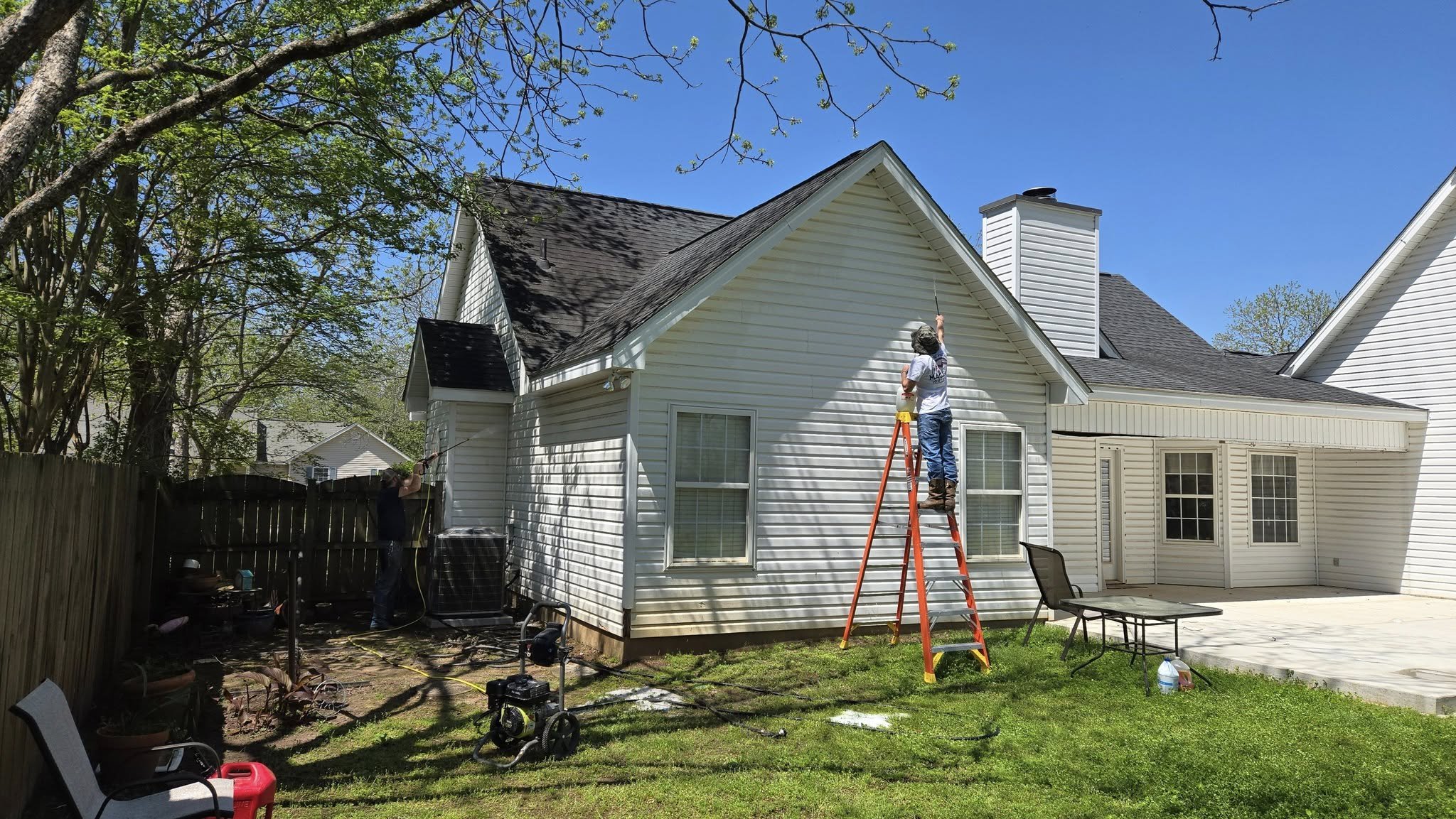 Two workers power wash the exterior of a white house with a ladder and nearby patio furniture on a sunny day.