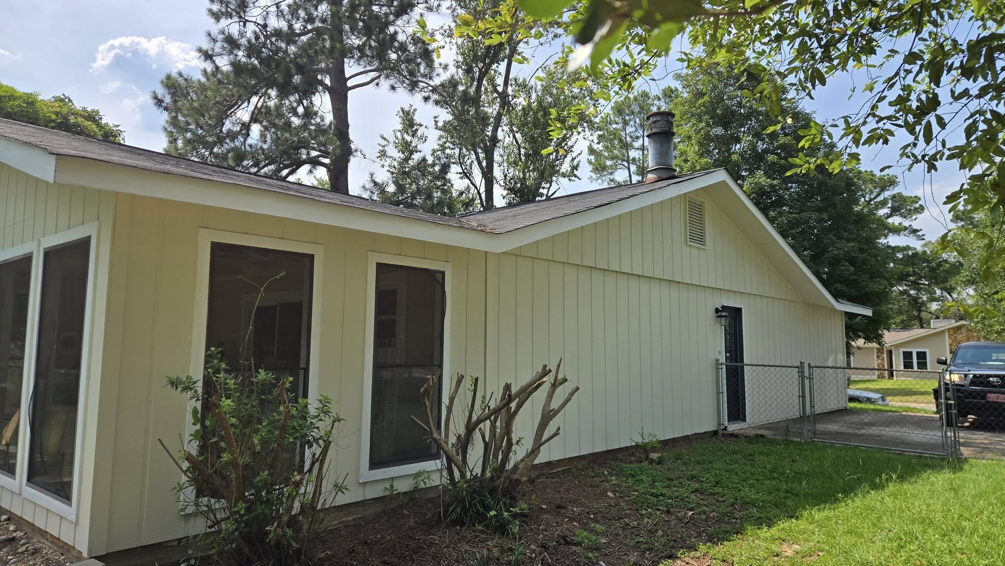 A yellow house with white trim, multiple windows, a black door, and a chain-link fence, with trees and a blue sky in the background.