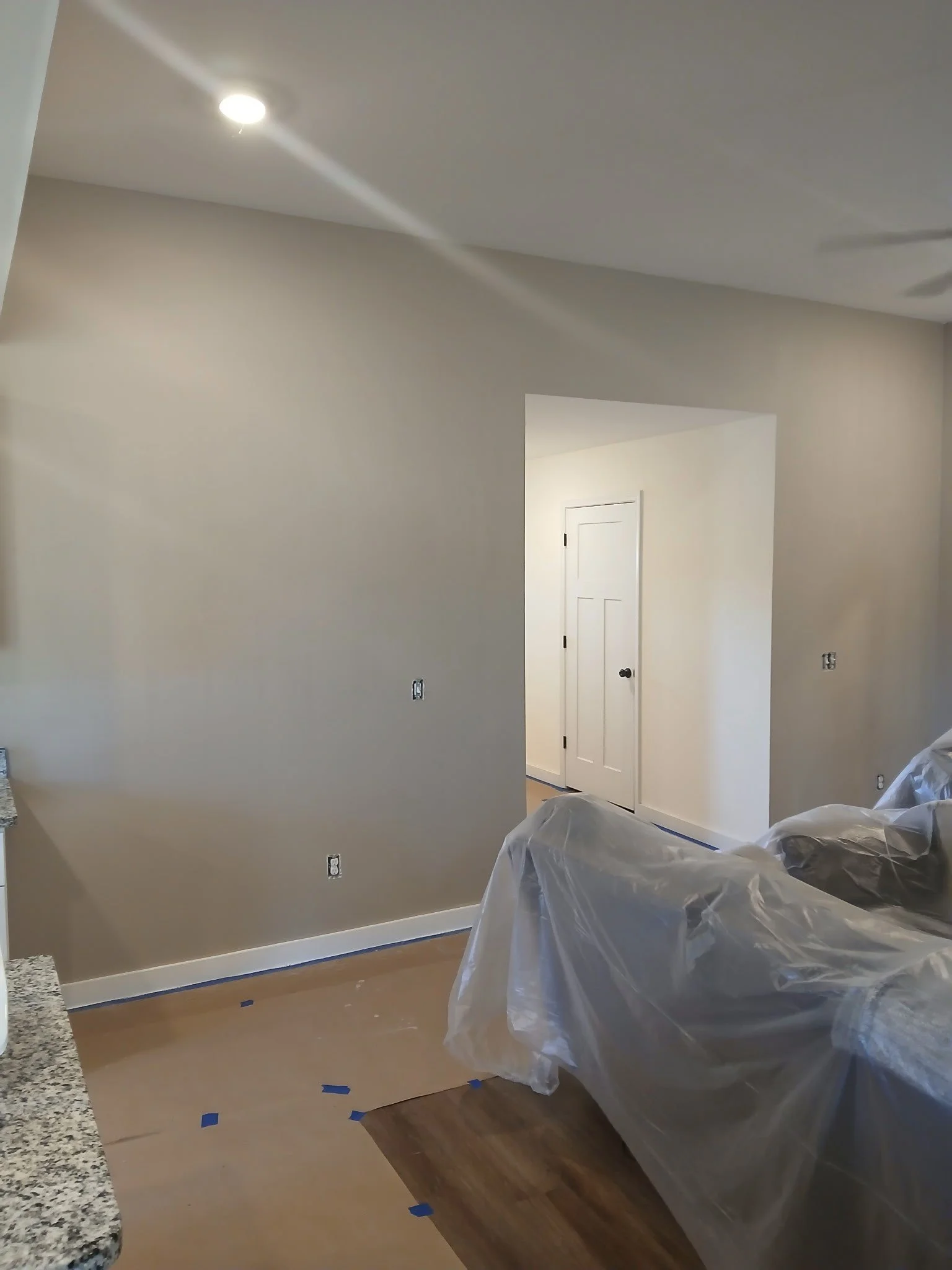 Interior of a home under renovation with beige painted walls, a doorway, and a plastic-covered couch on a hardwood floor, with painter's tape on the floor.