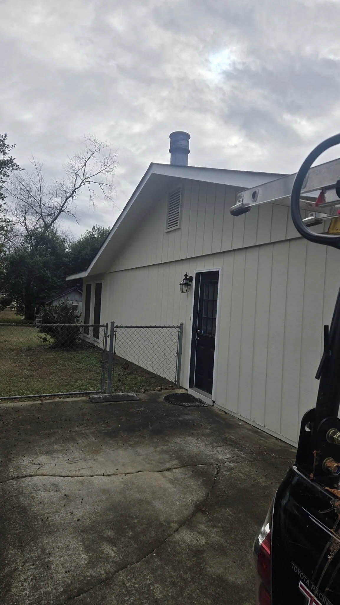 A freshly painted white house with a black door, a small outdoor light, and a concrete patio. A chain-link fence runs along the side, and trees are visible in the background under a cloudy sky.