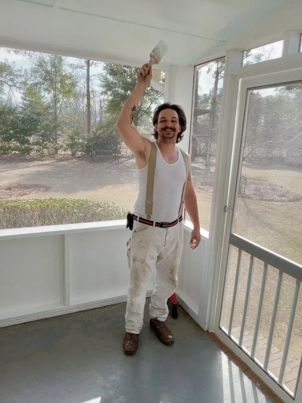 Man with dark hair, beard, wearing work clothes and suspenders, holding a dollar bill in the air inside a screened porch, smiling.