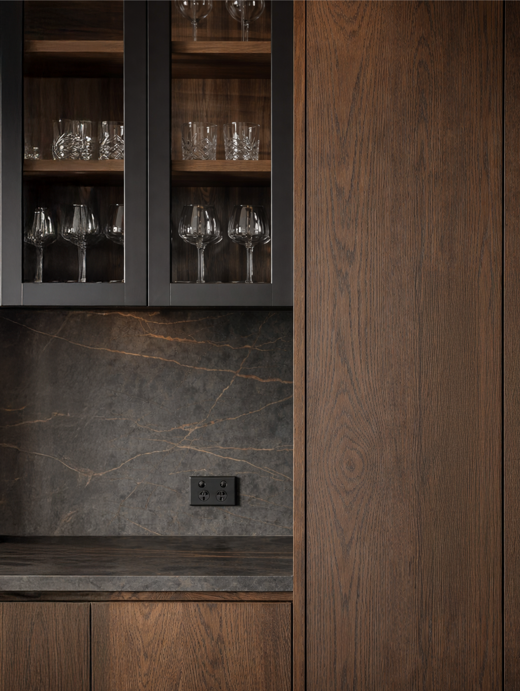 A kitchen cabinet with dark wooden finish, glass doors revealing wine glasses inside, and a black marble countertop with light veining. There is an electrical outlet on the wall.