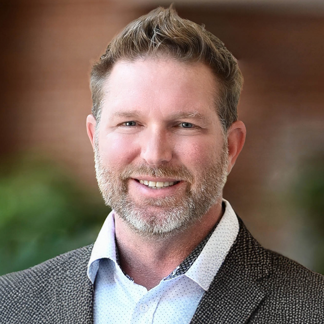 Close-up portrait of a smiling man, Don O'Shields, an inspector with Providence Inspections in Atlanta, with light brown hair and beard, wearing a white shirt and a dark blazer, with a blurred outdoor background.
