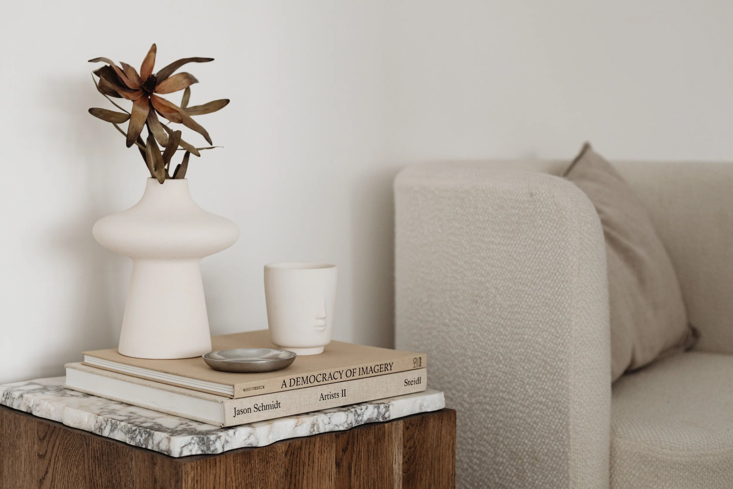 Table with a marble top holding stacked books and decorative items, including a white vase with dried flowers, a small face-shaped ceramic cup, and a silver dish, adjacent to a light-colored sofa.
