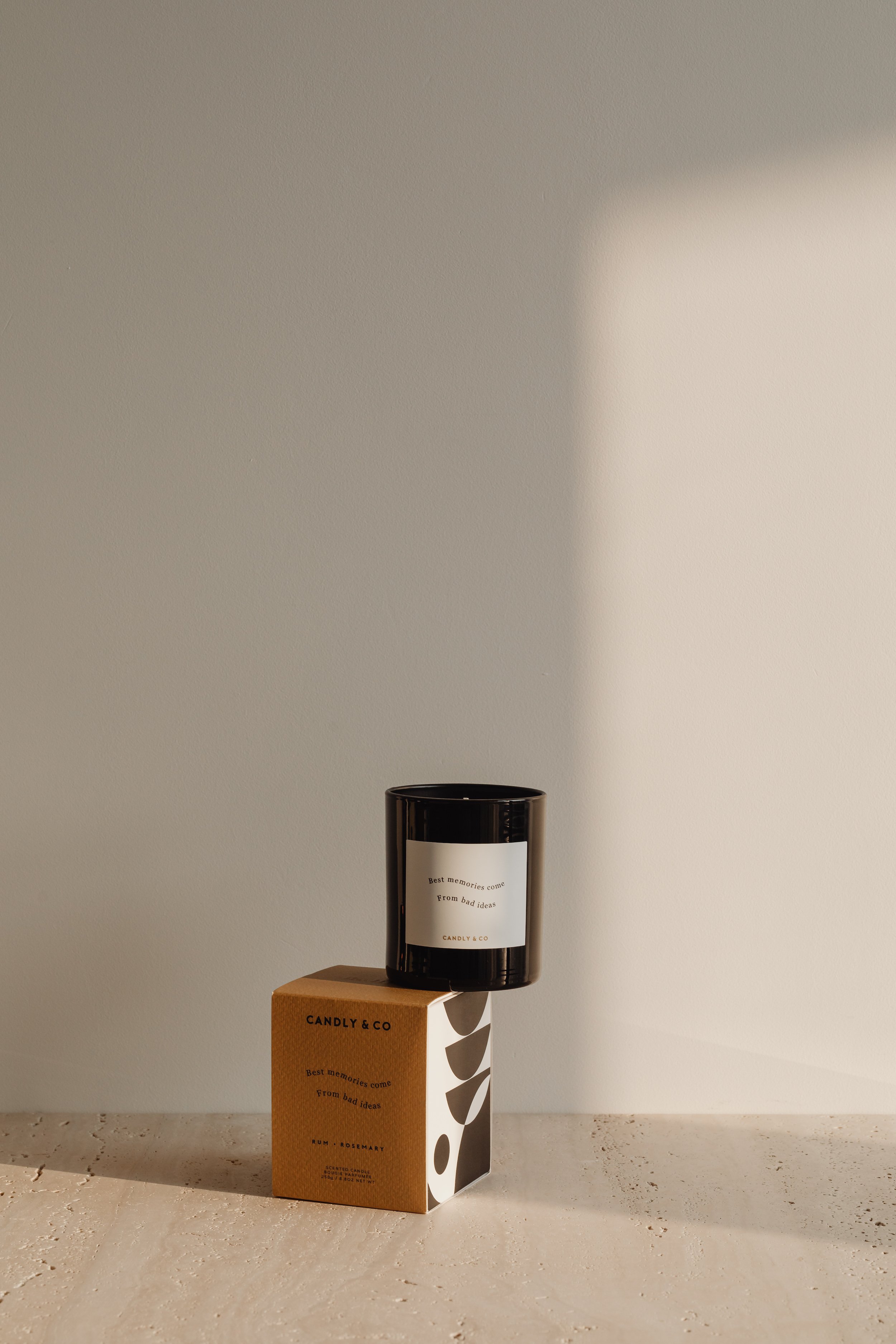 A black scented candle container on top of a small decorative box with a plant pattern. The box and candle have labels with the text 'Best memories come from bad ideas. CANDLY & CO' and 'Rum + Rosemary'.