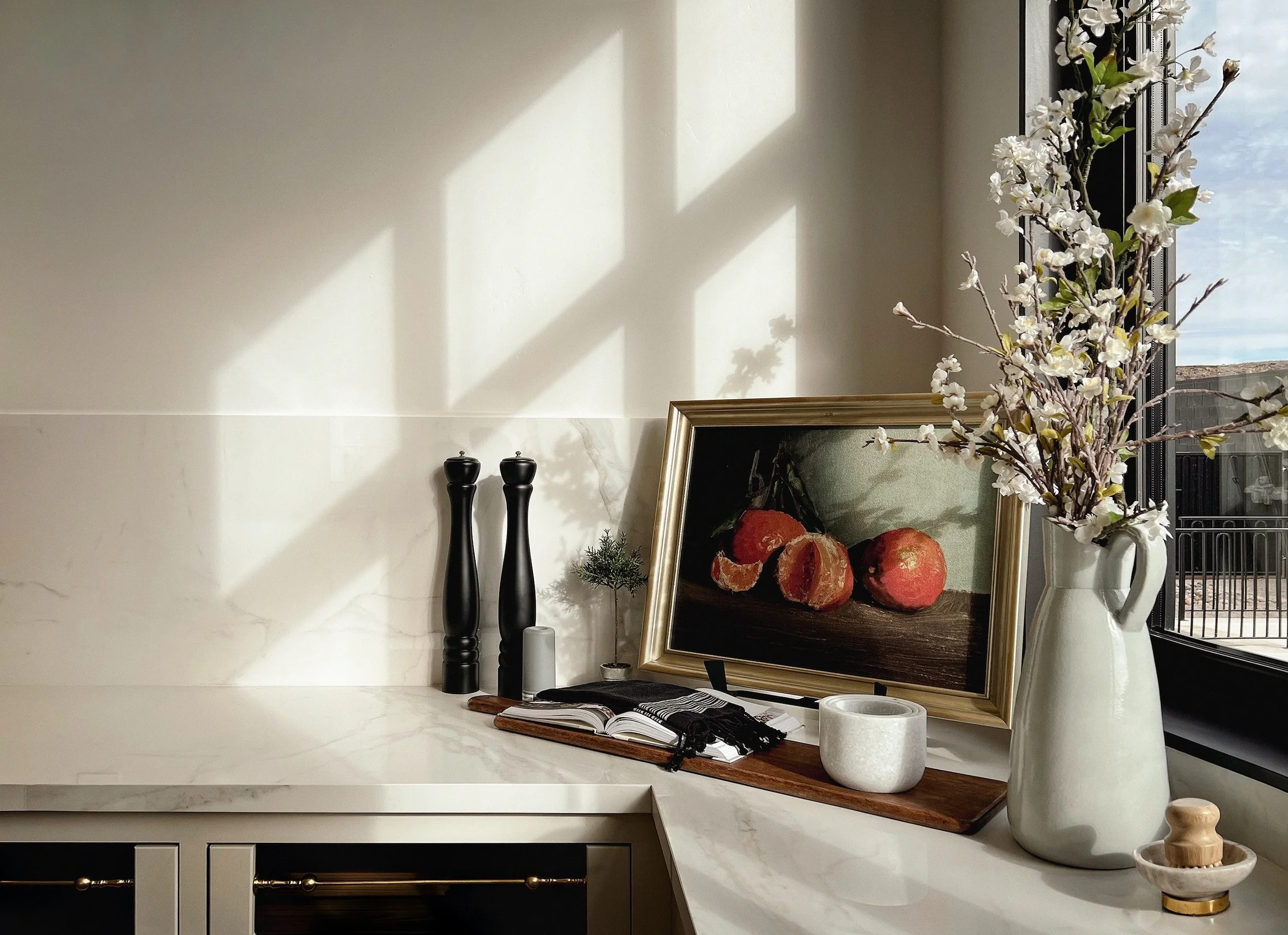 Decorative kitchen counter with black pepper and salt shakers, small potted plant, framed painting of oranges, open book, white vase with flowers, and a window with sunlight and view outside.