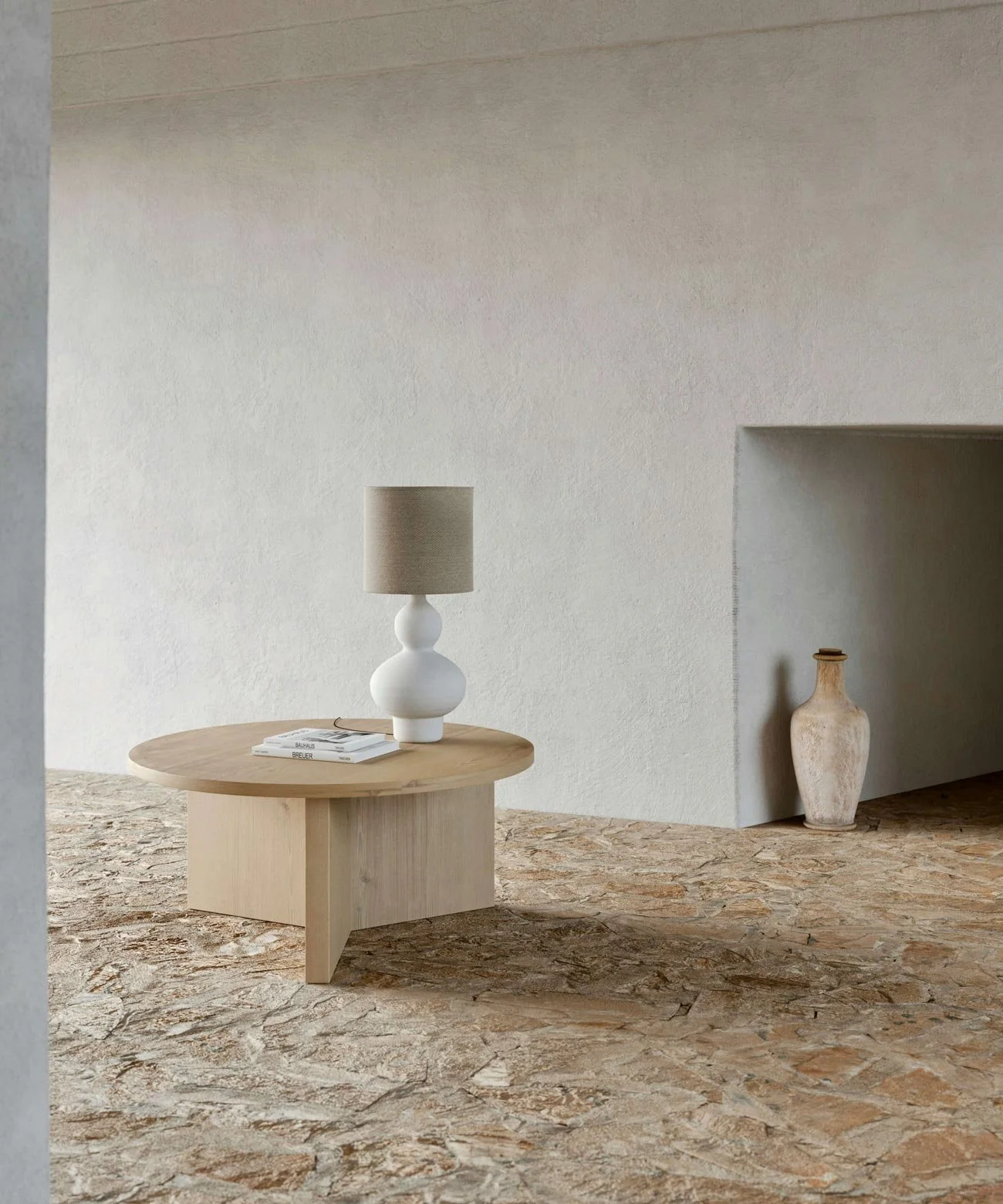 Minimalist interior with a round wooden side table, a white ceramic table lamp, a stack of books, and a large beige ceramic vase in a niche on the wall.
