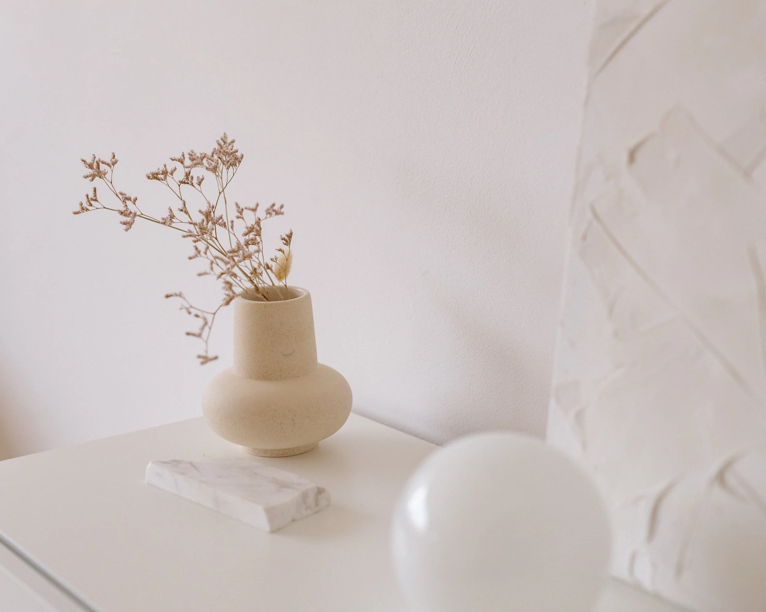 A beige vase with dried flowers on a white surface, next to a small white marble piece and a white decorative object with embossed patterns.