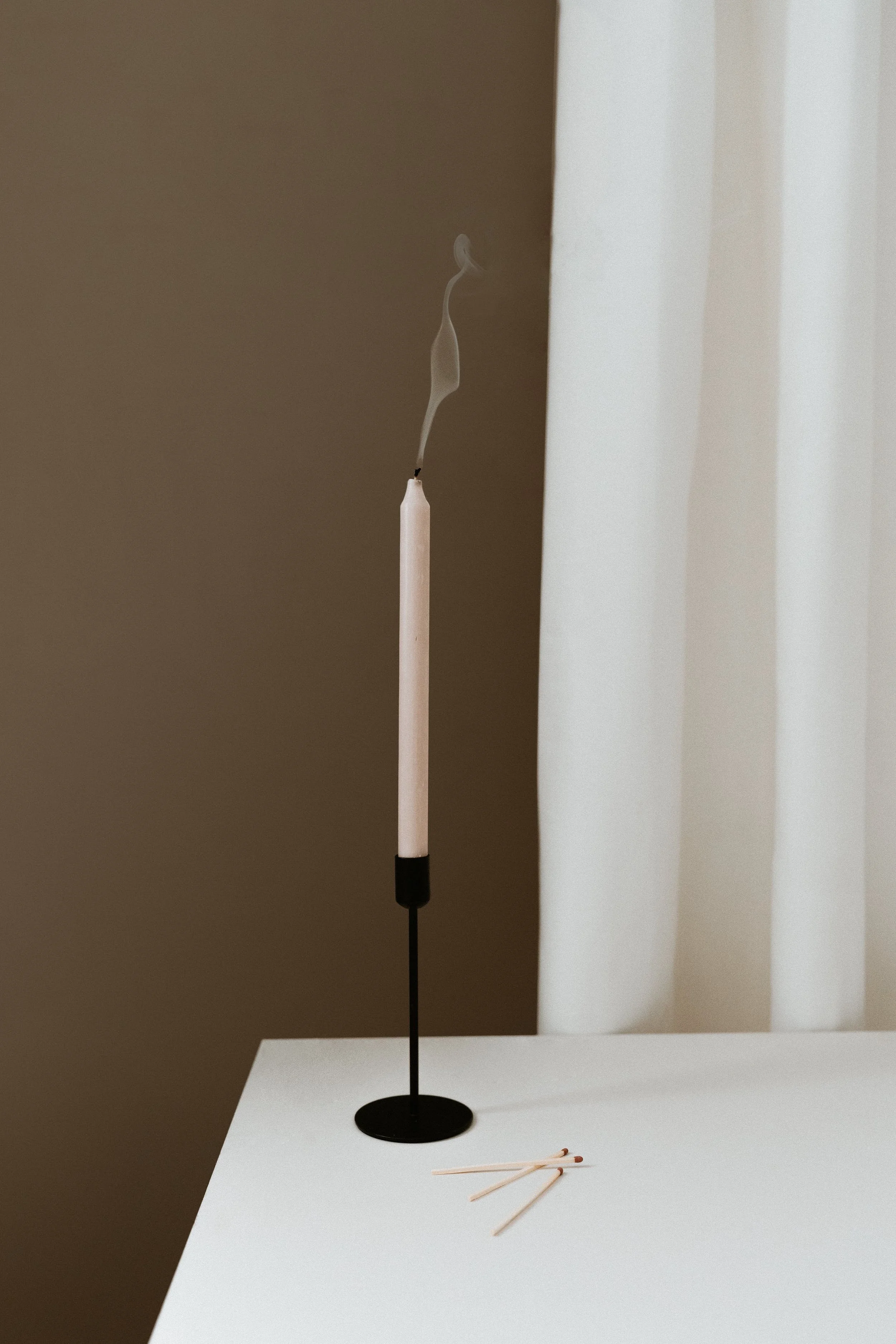 A lit white candle on a black stand with smoke rising and three matchsticks on a white surface.
