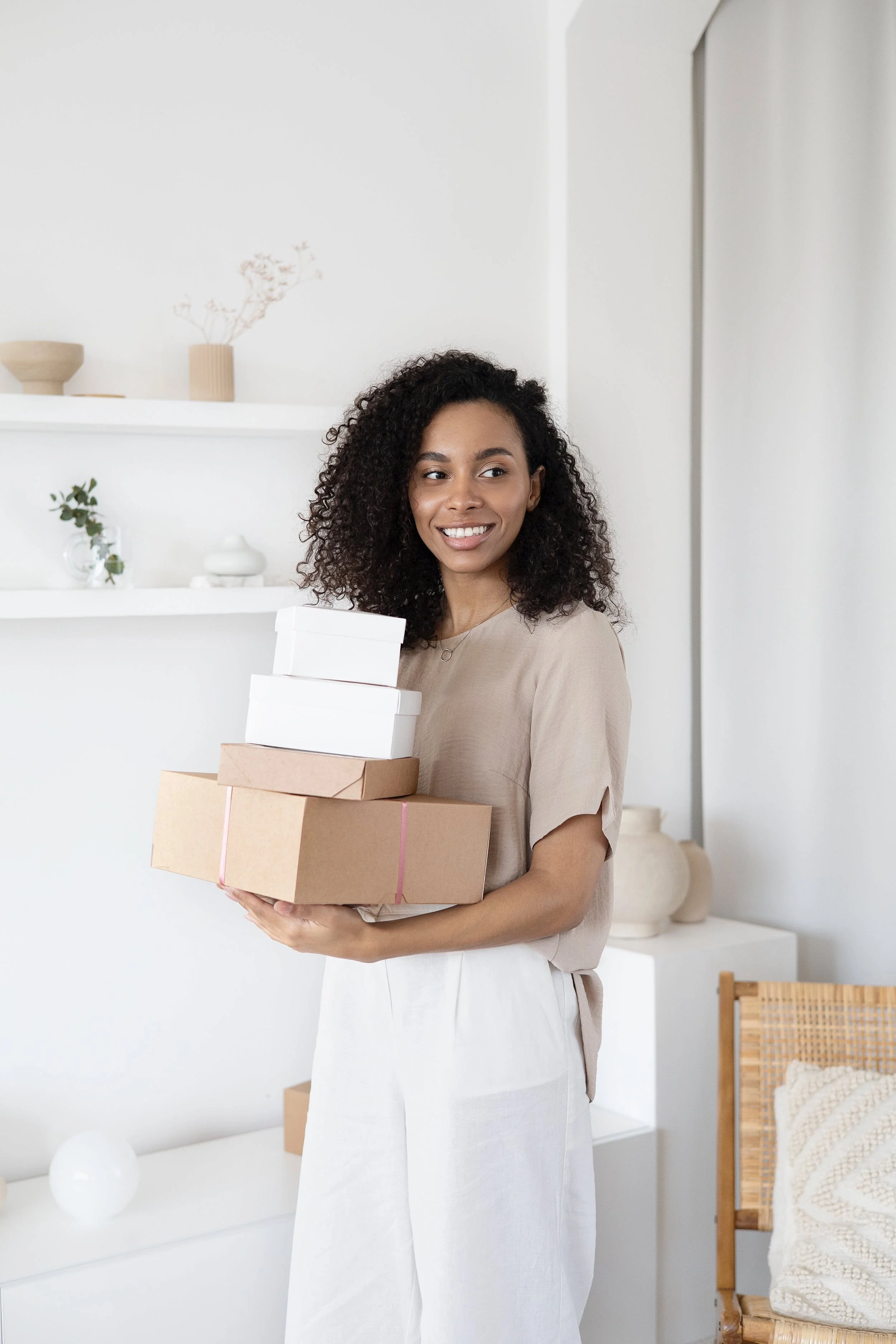 A smiling woman holding a stack of gift boxes in a minimalistic, white room with decorative shelves and vases.