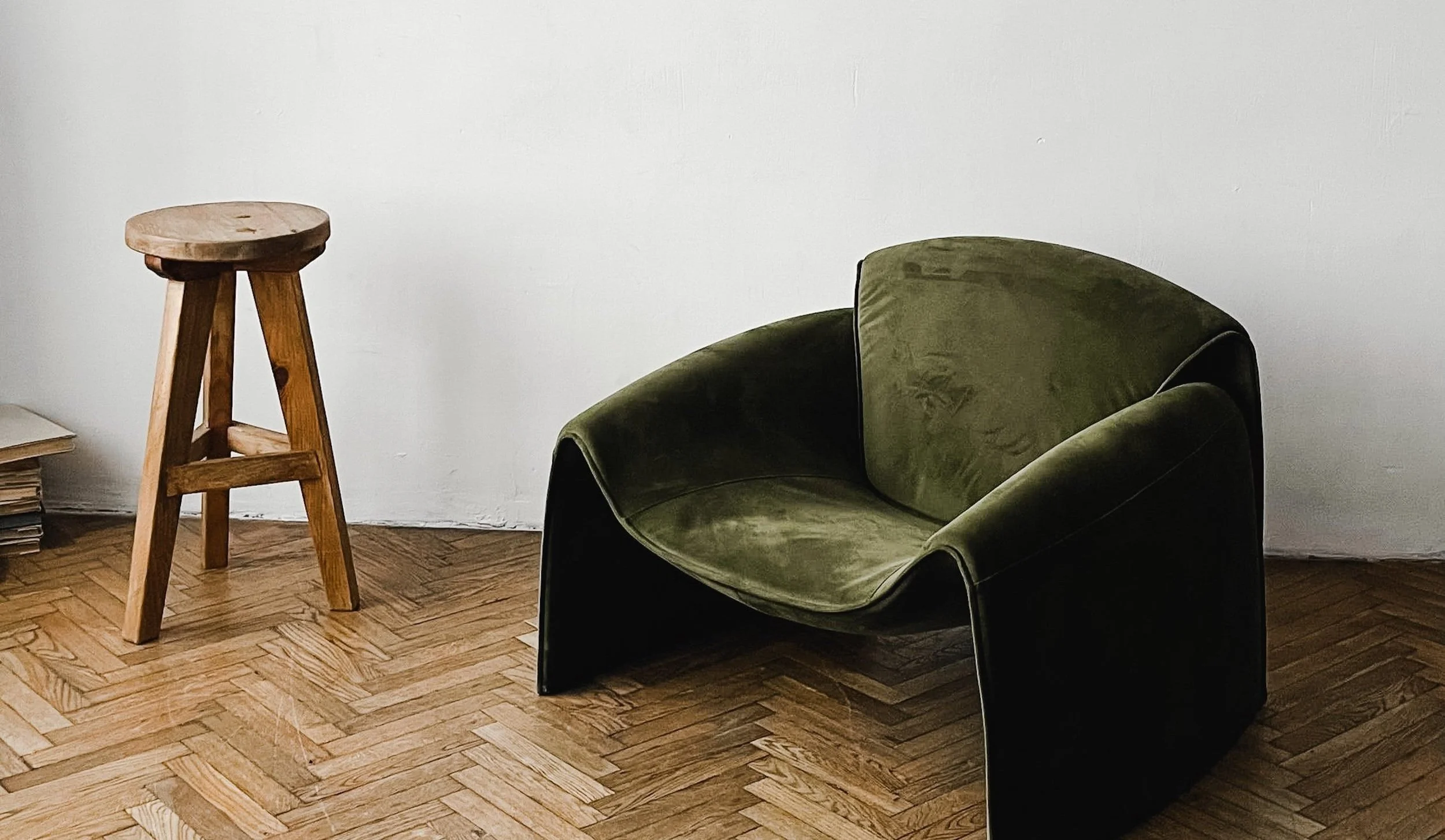 A green upholstered armchair with black sides next to a wooden stool in a room with a white wall and wooden floor.