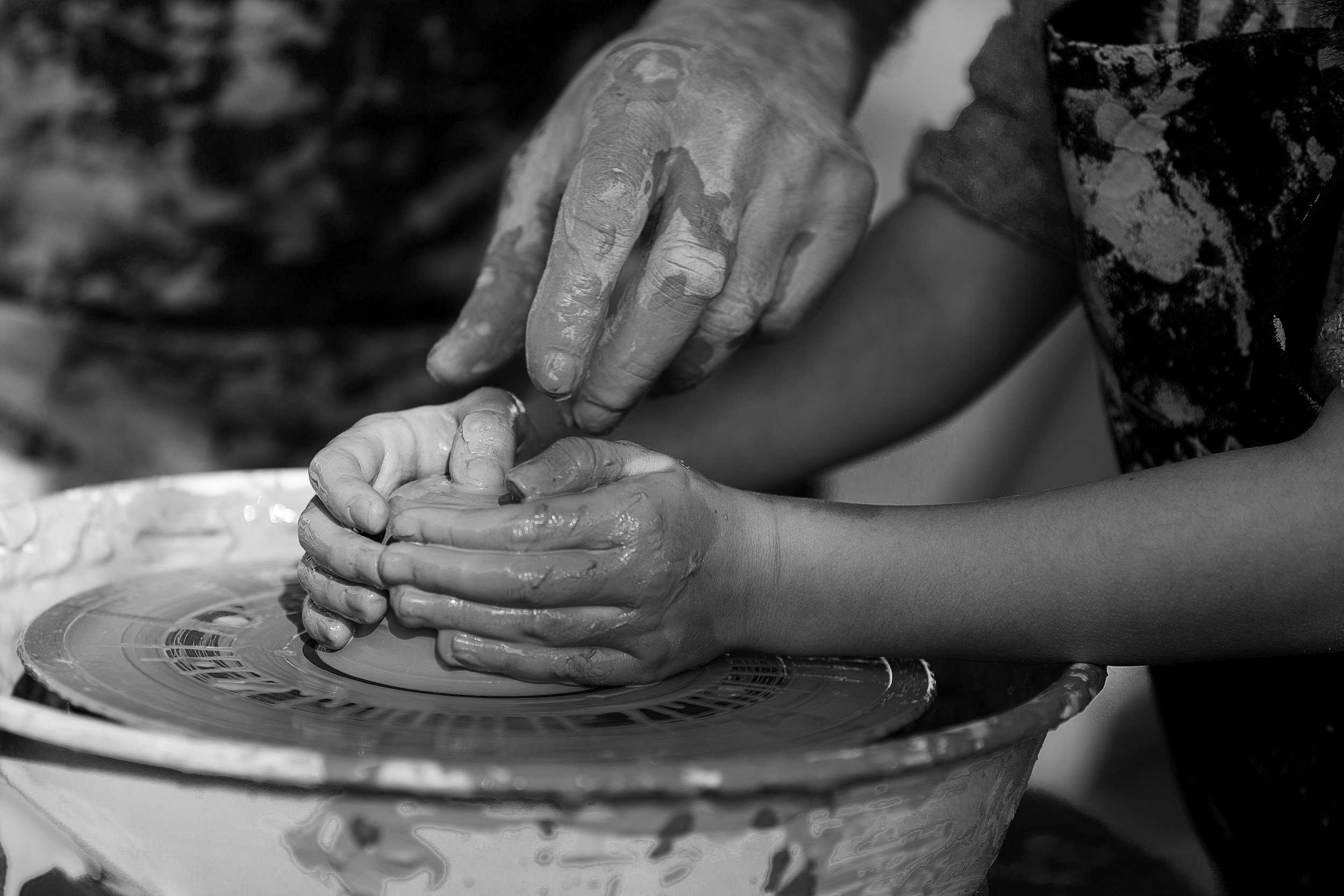 Close-up of two people engaging in pottery-making; one person guides the other's hands on a spinning pottery wheel, both hands covered in clay.