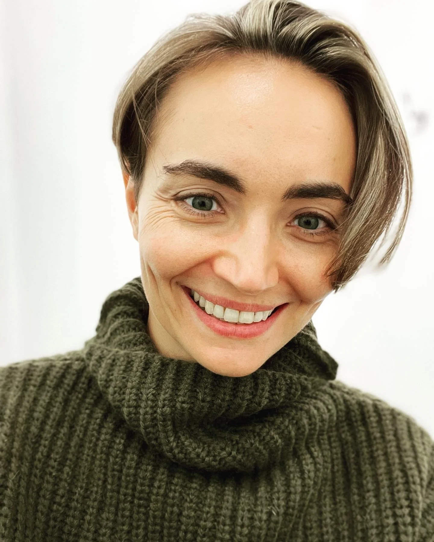 A smiling woman with short brown hair and blue eyes, wearing a dark green chunky knit turtleneck sweater, in front of a plain white background.