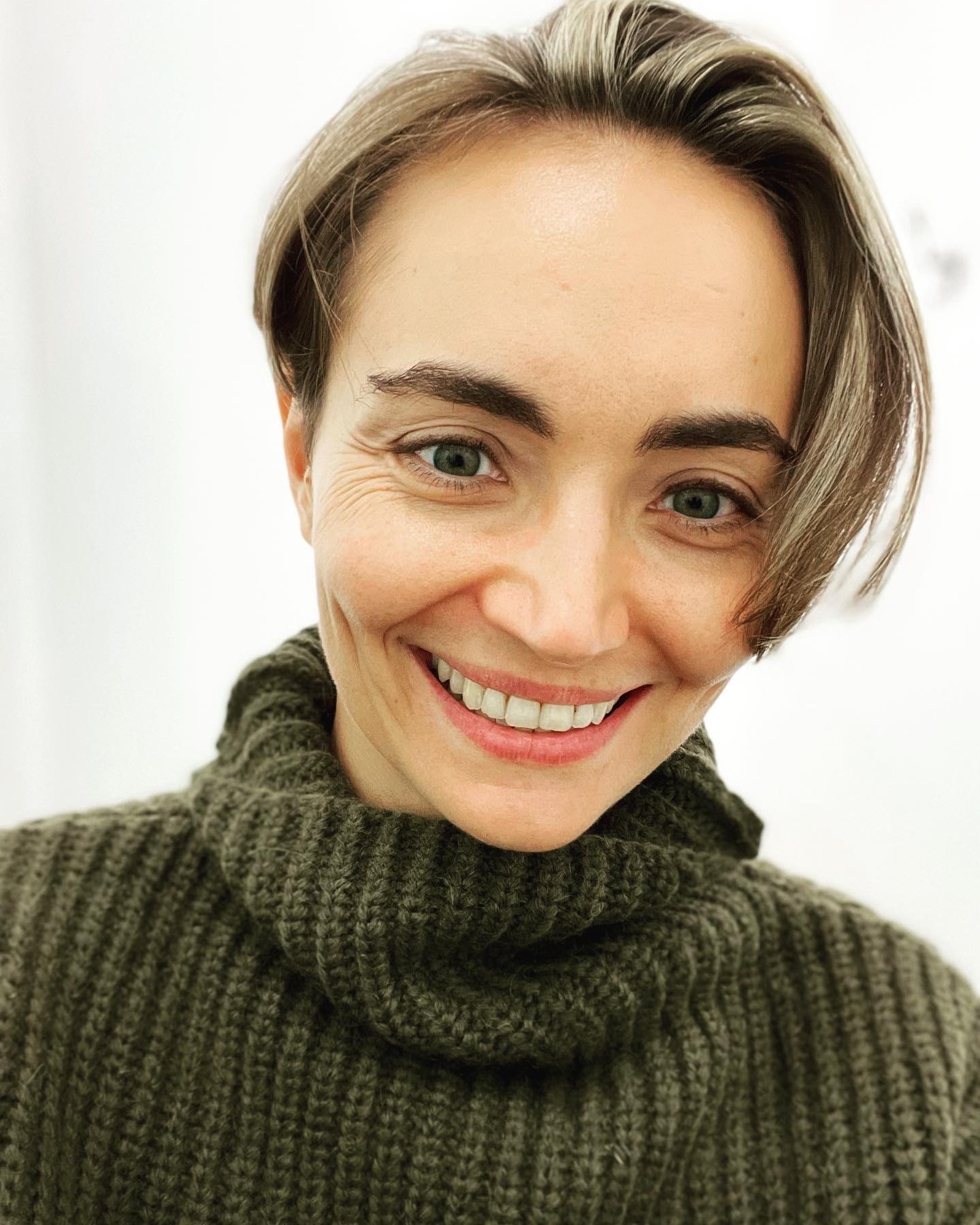 Close-up of a woman with short brown hair, blue eyes, smiling, wearing a green knitted turtleneck sweater.