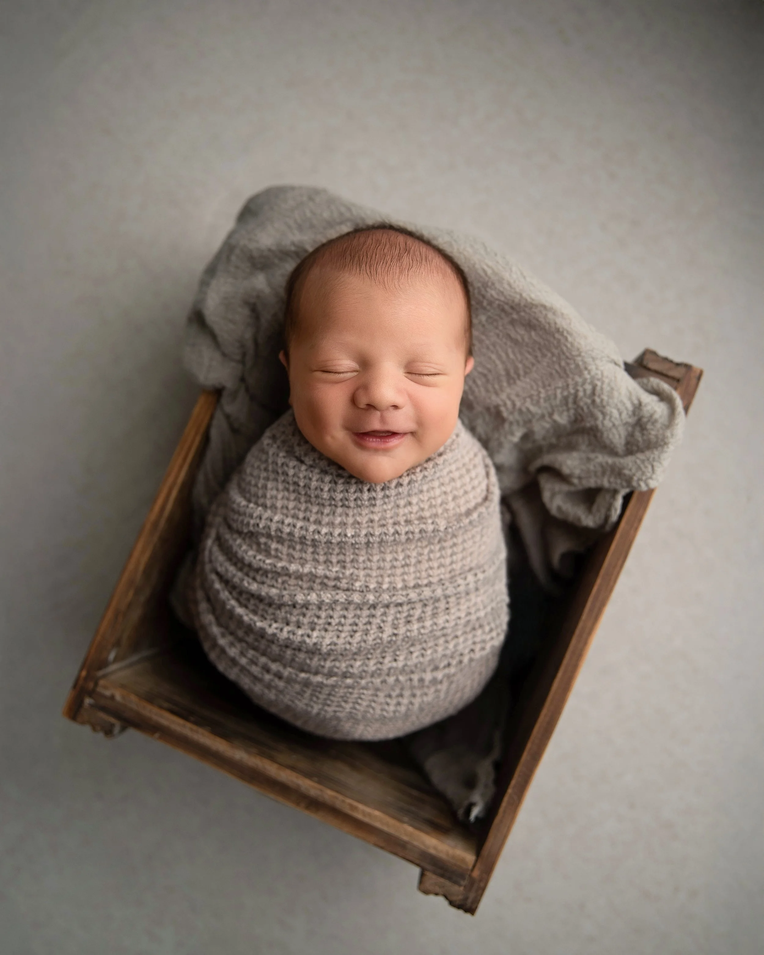 A smiling baby wrapped in a beige knit blanket, lying in a small wooden crate with a gray blanket fall behind him