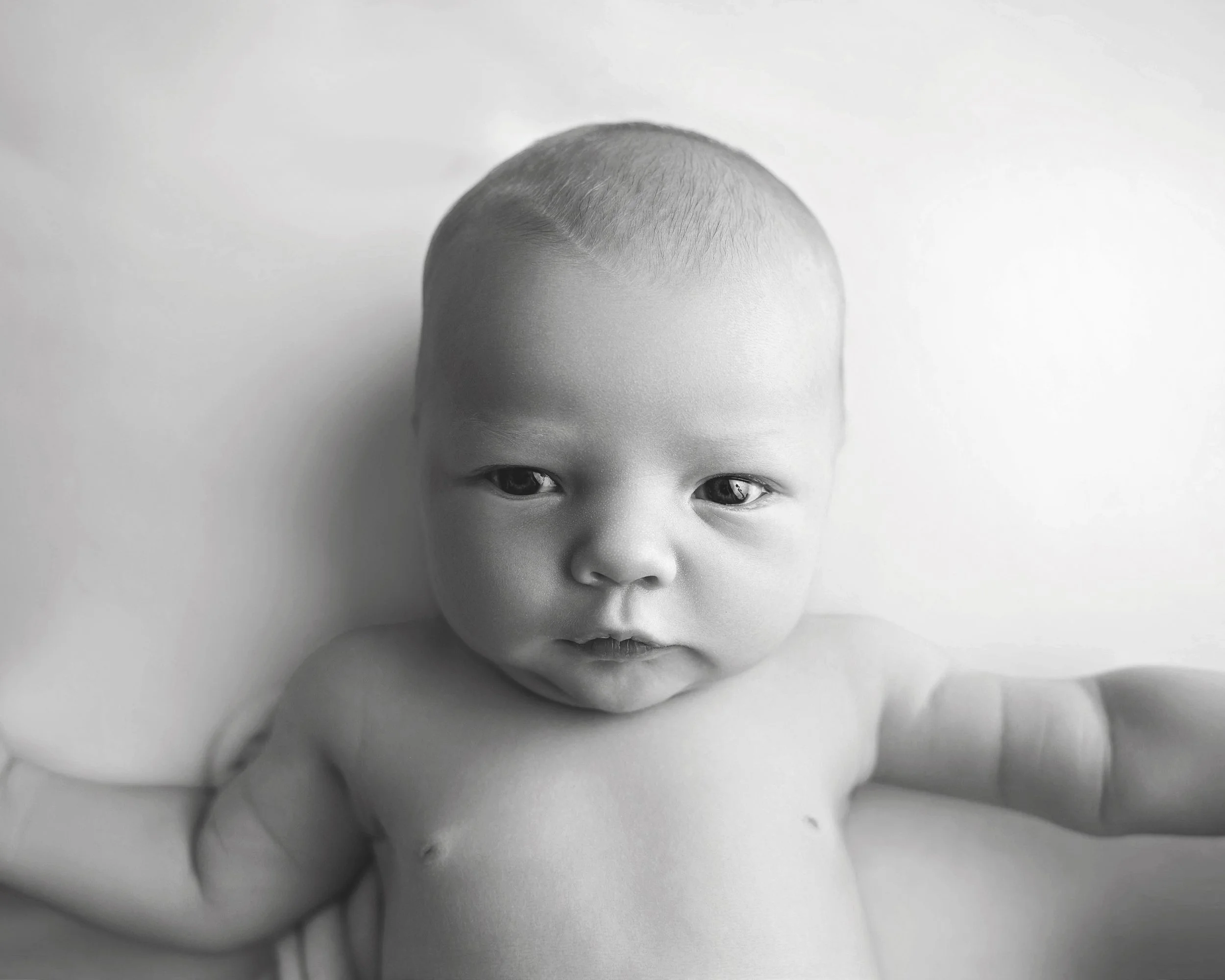 Black and white photo of a baby with light skin and short hair looking directly at the camera, lying on a flat surface with arms extended.