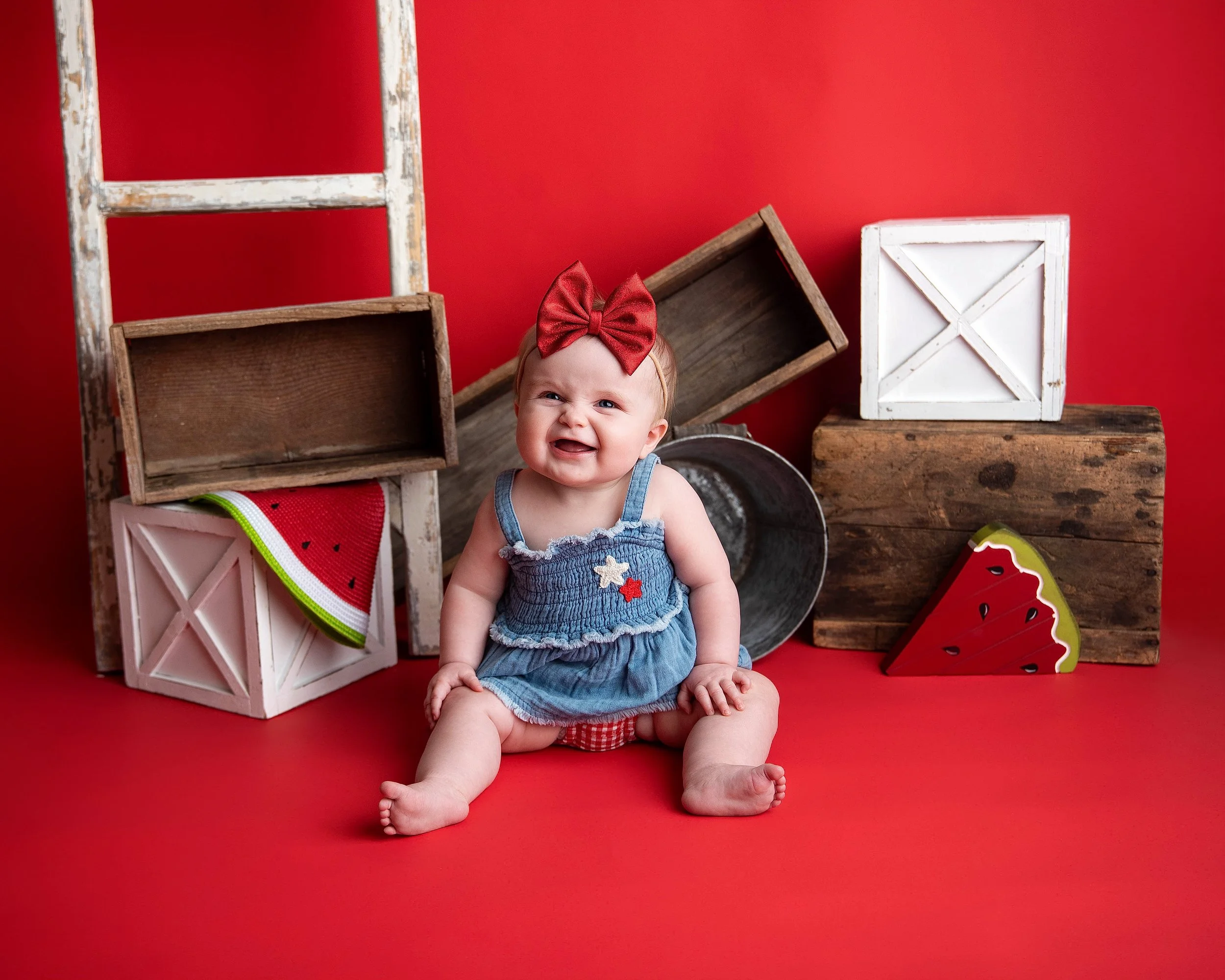 A baby girl wearing a blue dress with a red bow headband, sitting on a red background with wooden crates and watermelon decorations around her.