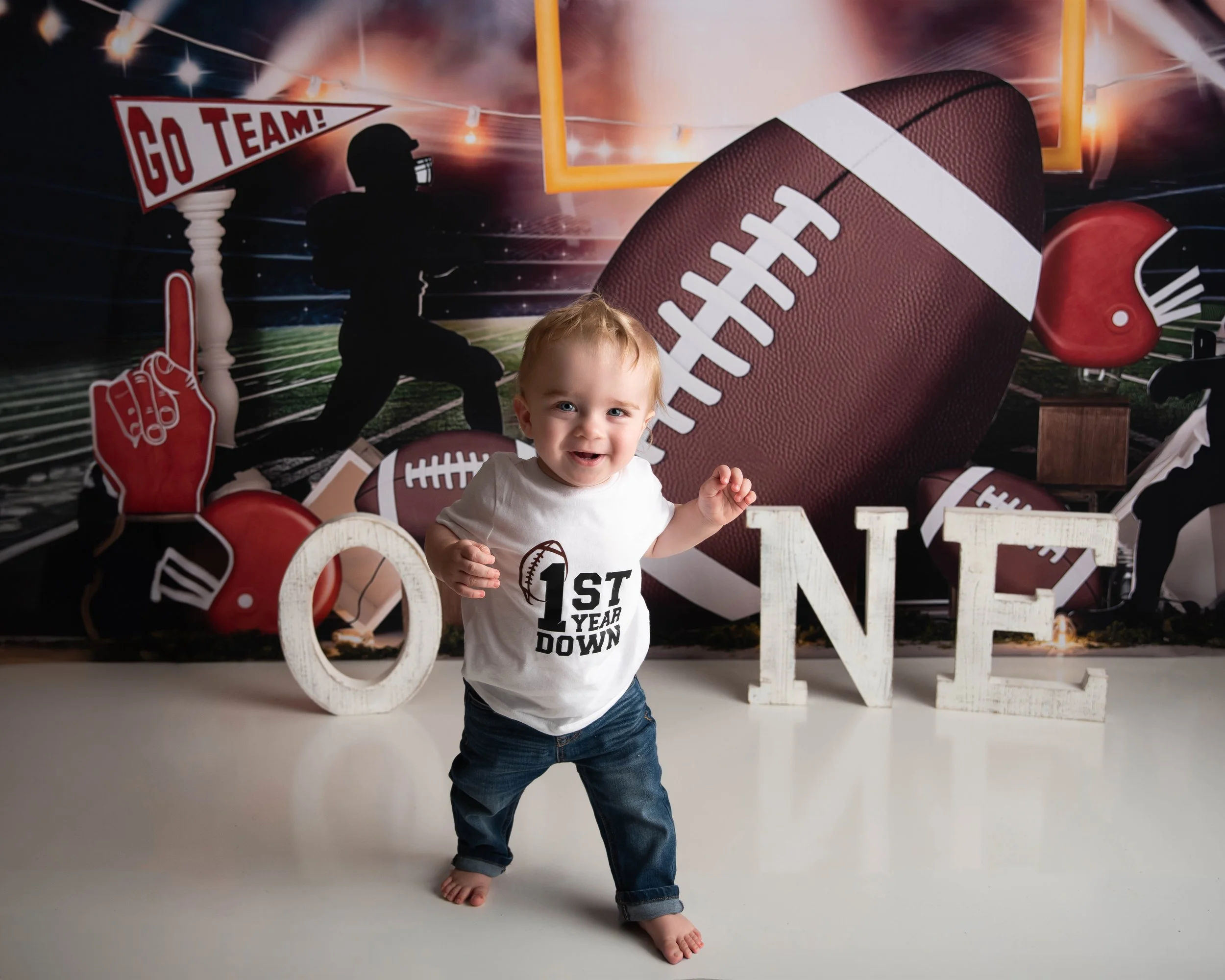 A smiling toddler standing on a white floor in front of a football-themed backdrop, with large white letters spelling out 'ONE'. The toddler is wearing a white shirt that says '1st Year Down' and jeans, with one foot slightly raised and arms bent.
