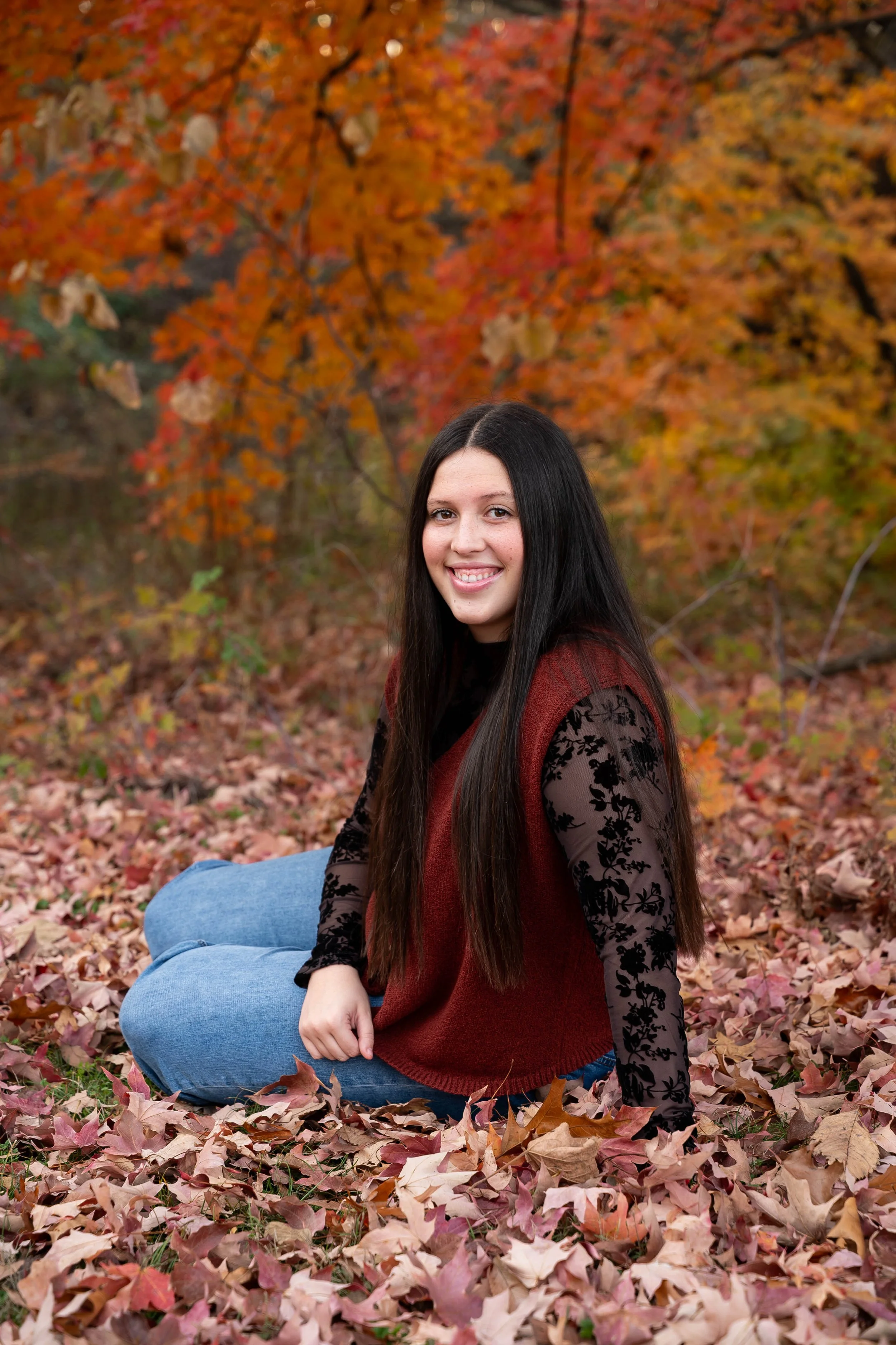 A young woman with long dark hair sitting on a bed of fallen autumn leaves surrounded by trees with orange and yellow fall foliage, smiling at the camera.