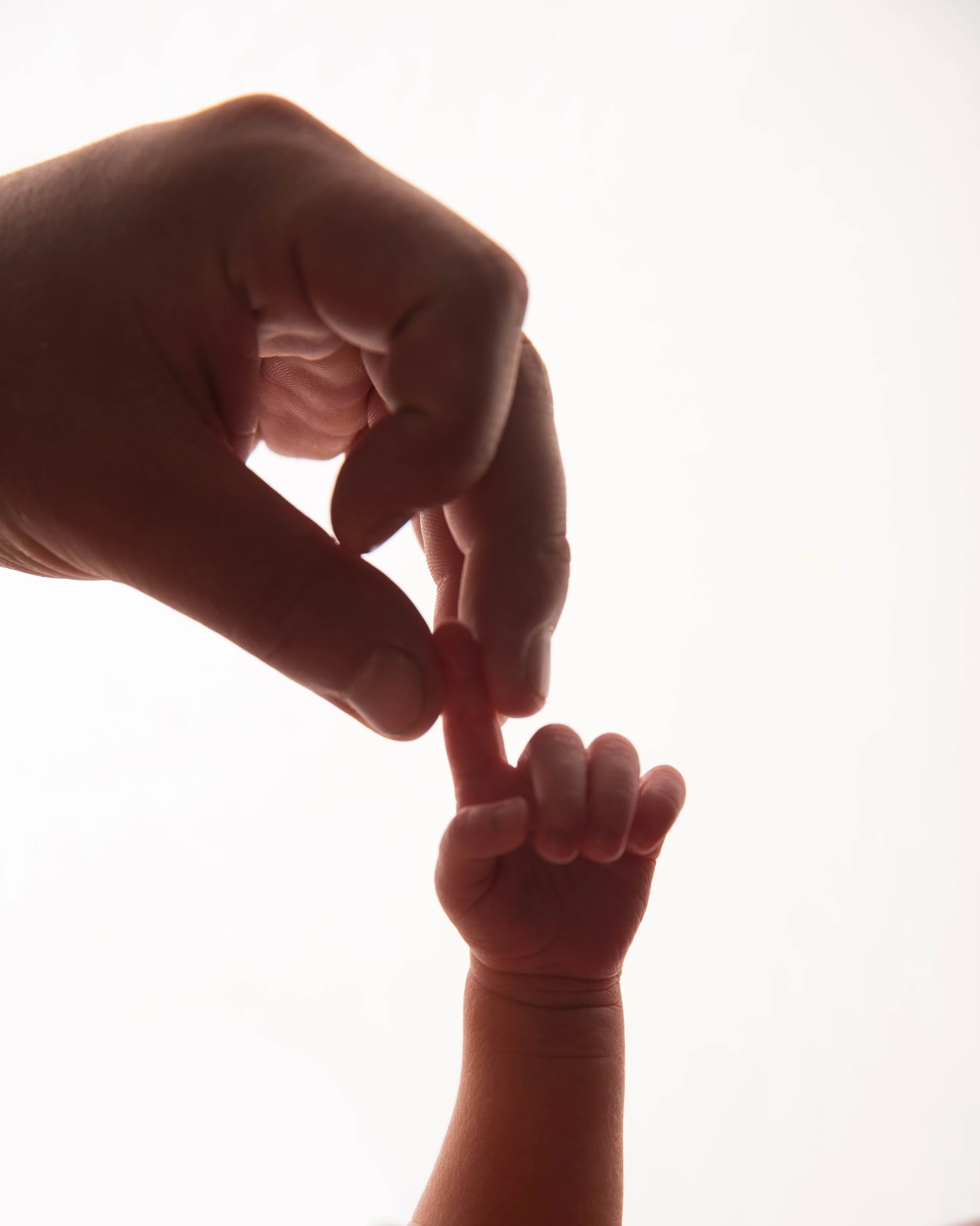 Close-up of a person's hand holding the small finger of a child's hand, with the person gently touching the child's finger against a bright, white background.