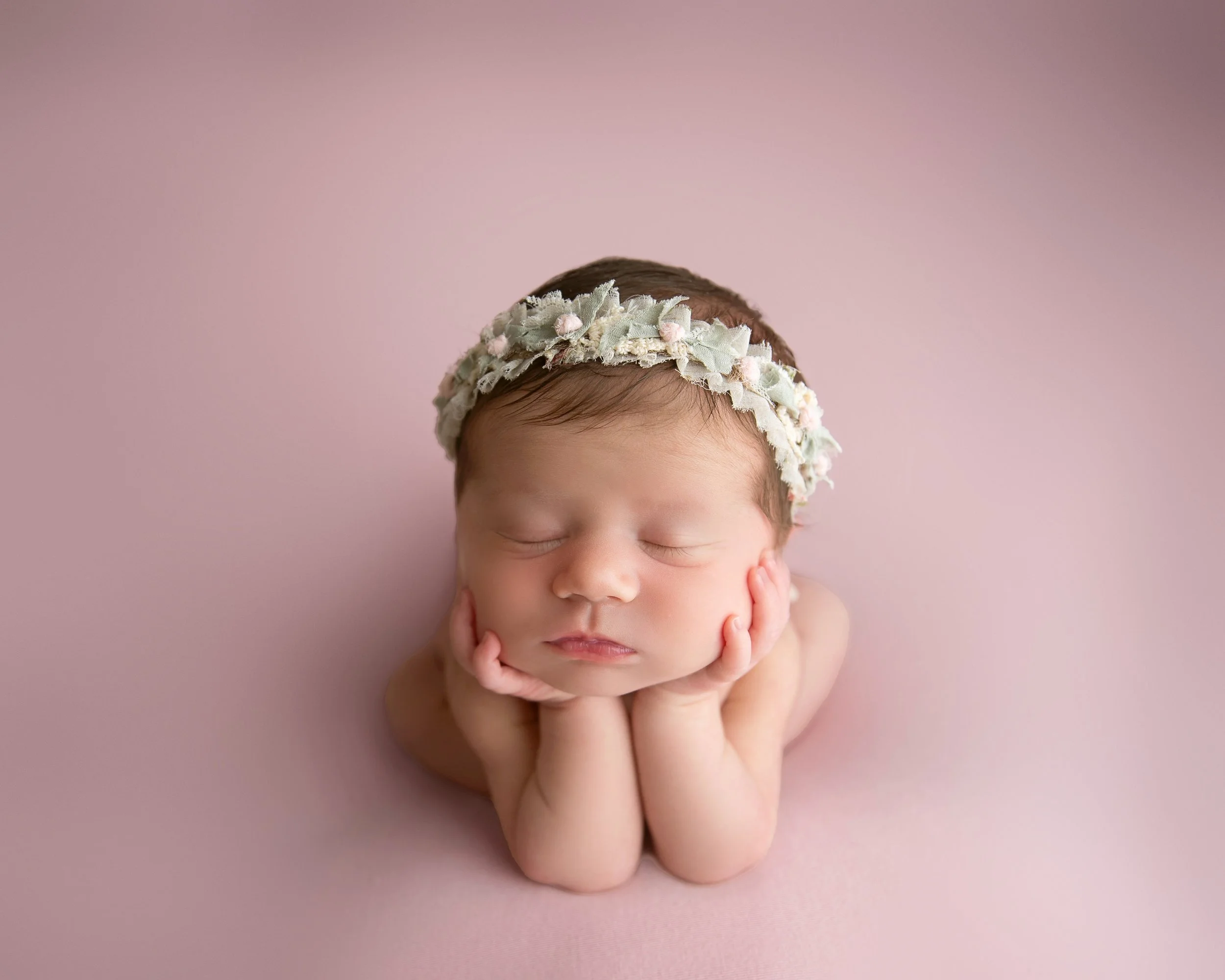 A sleeping newborn baby with a floral headband, resting the chin on hands, on a pink background.