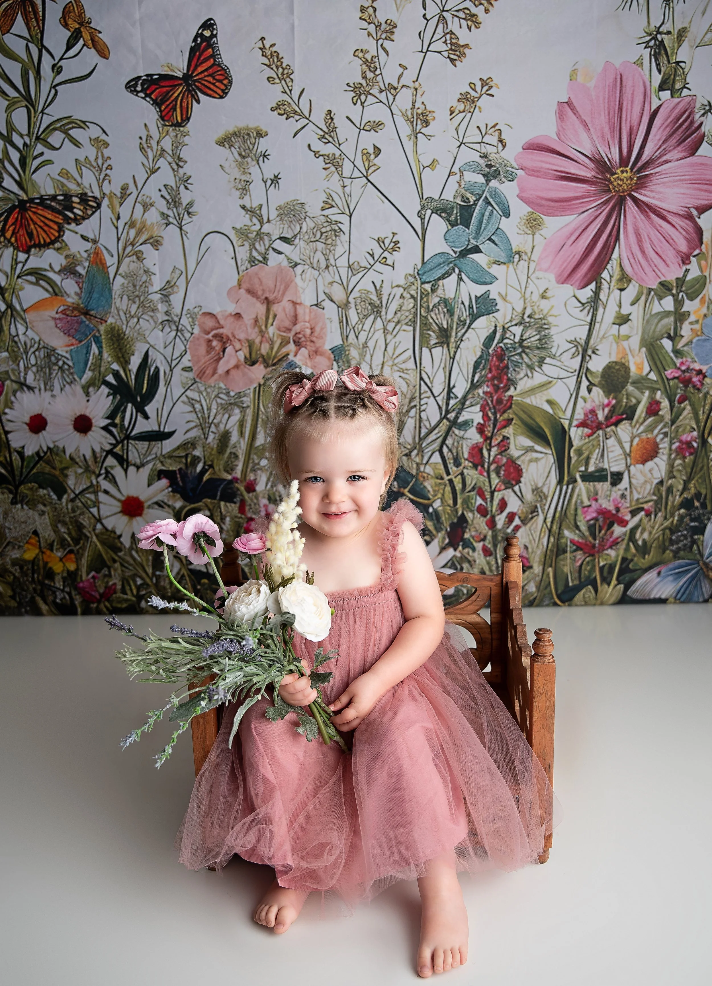 A young girl sitting on a small wooden bench, wearing a pink dress with a tulle skirt and pink ribbon hair bows, holding a bouquet of flowers, with a vibrant floral and butterfly mural background.
