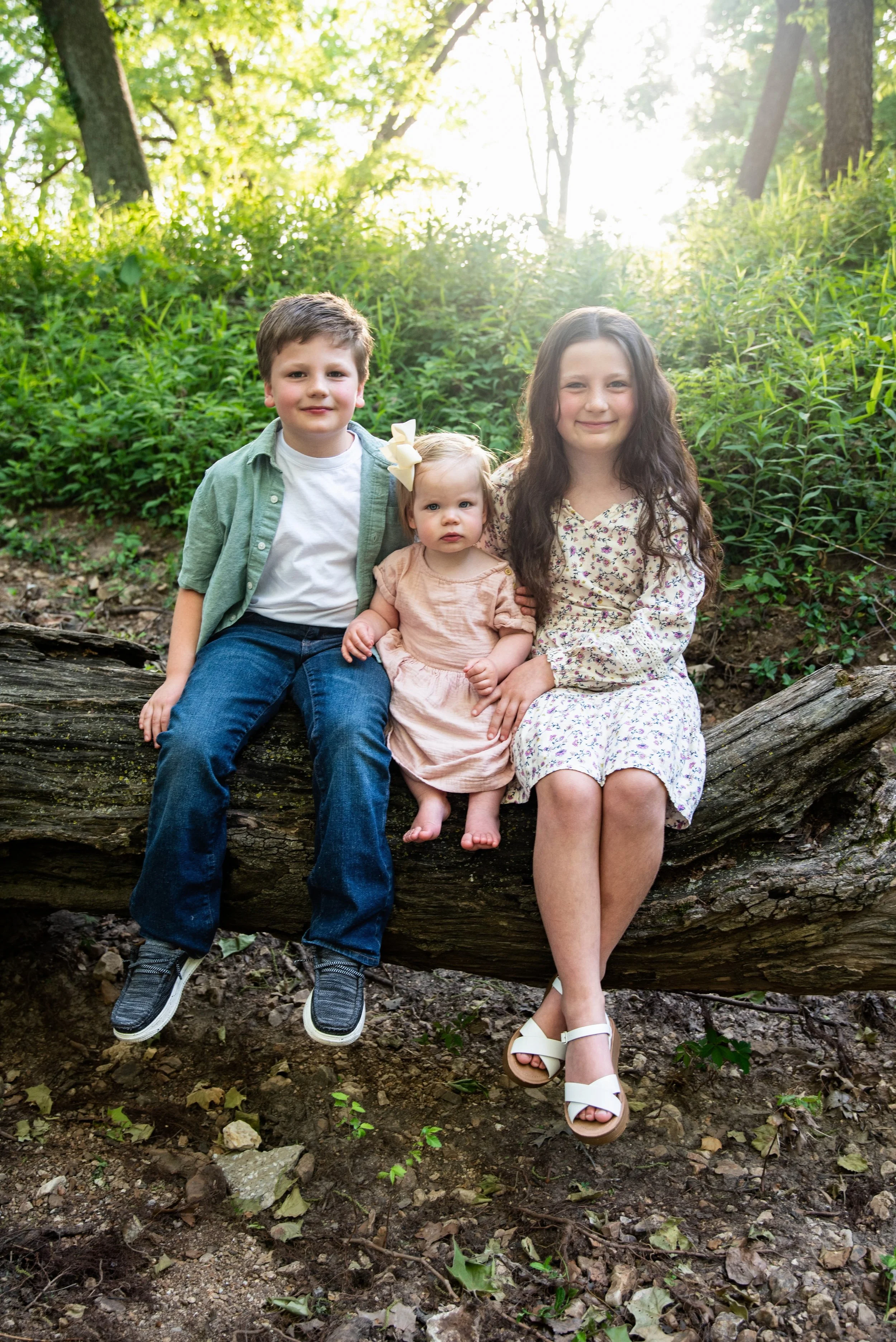 Three children, a boy, a girl, and a toddler girl, sitting on a large fallen tree in a forest with sunlight filtering through the trees.