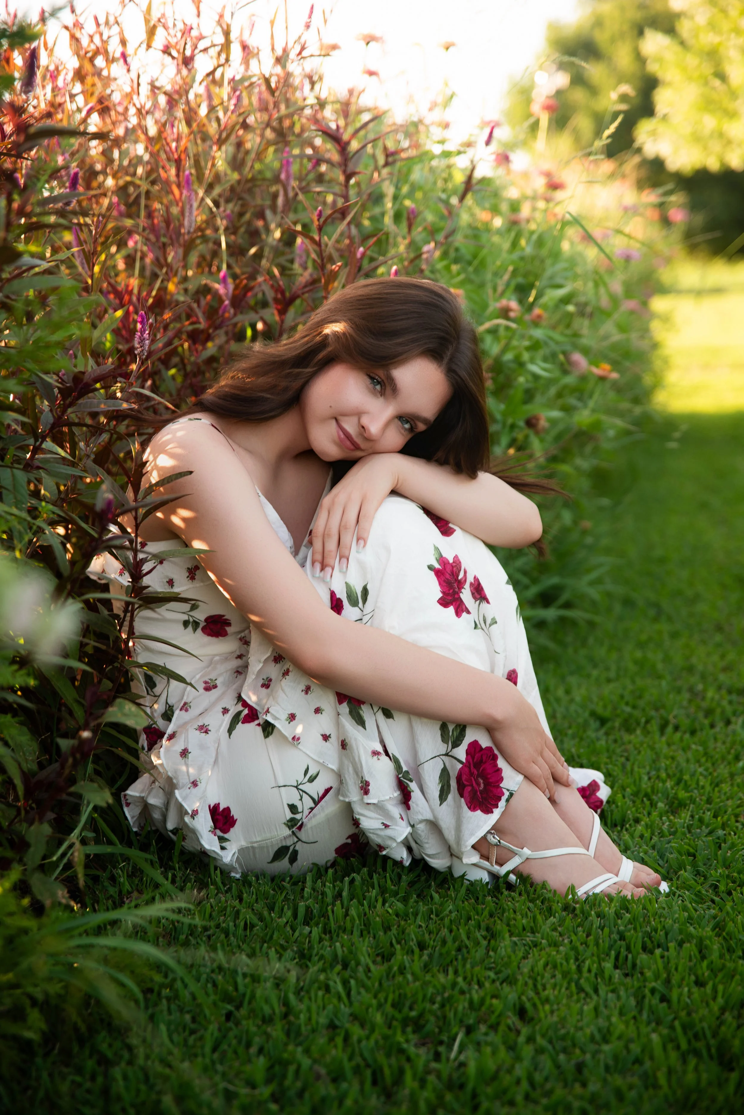 A young woman with long brown hair, sitting on green grass beside a flowering bush with pink and purple flowers, smiling gently at the camera during golden hour.