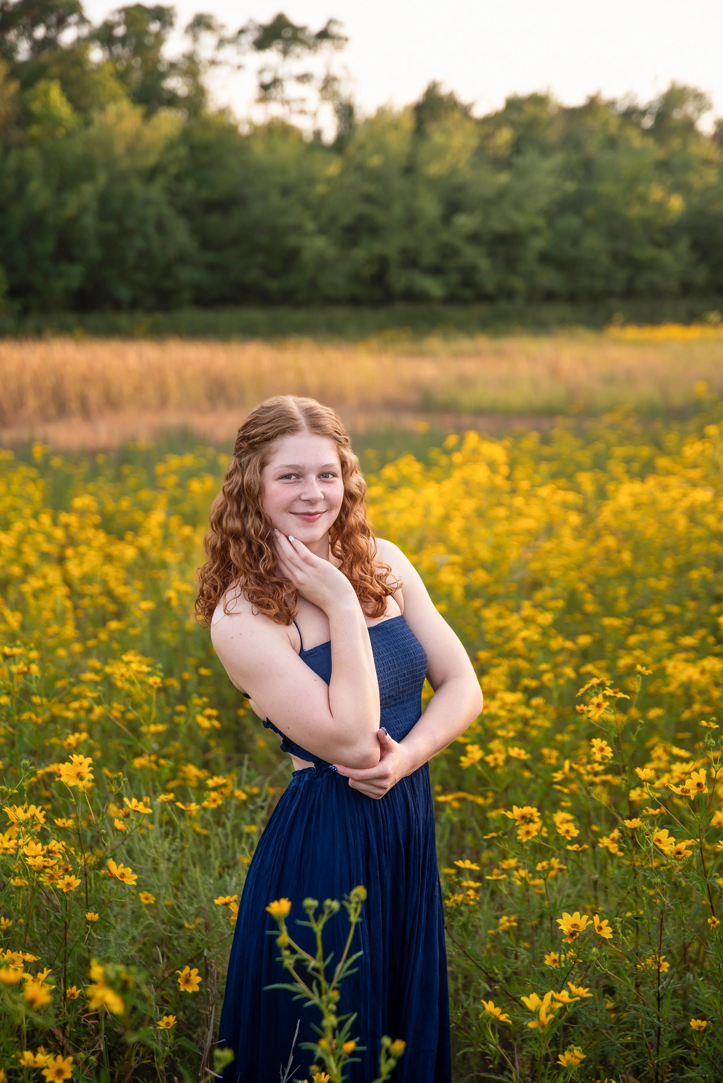 A young woman with curly red hair in a blue dress standing in a field of yellow flowers, smiling softly, with green trees in the background during sunset.