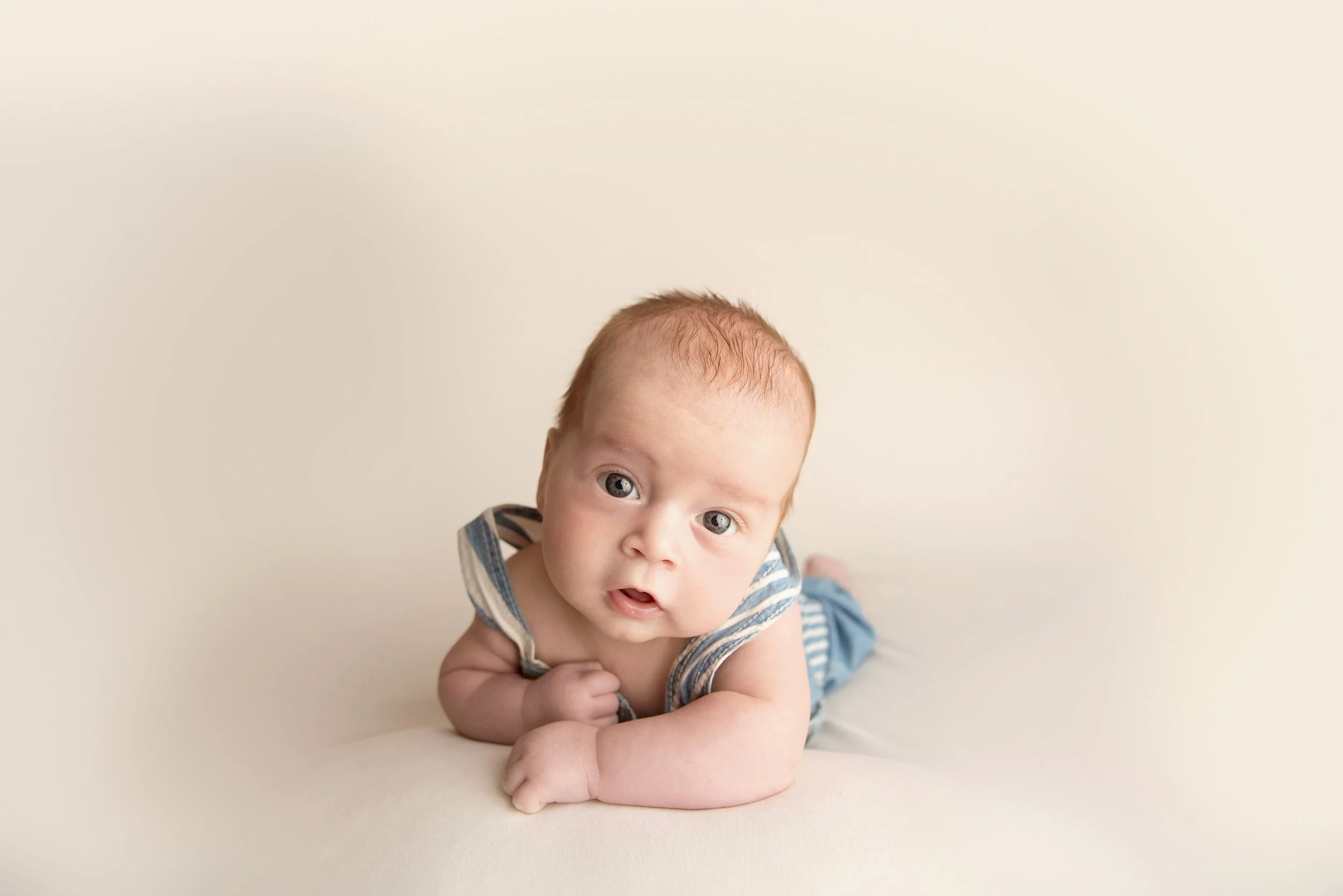 A baby with light skin and blue eyes, lying on his stomach on a cream-colored surface, wearing a striped blue and white sleeveless shirt and blue pants, looking directly at the camera.