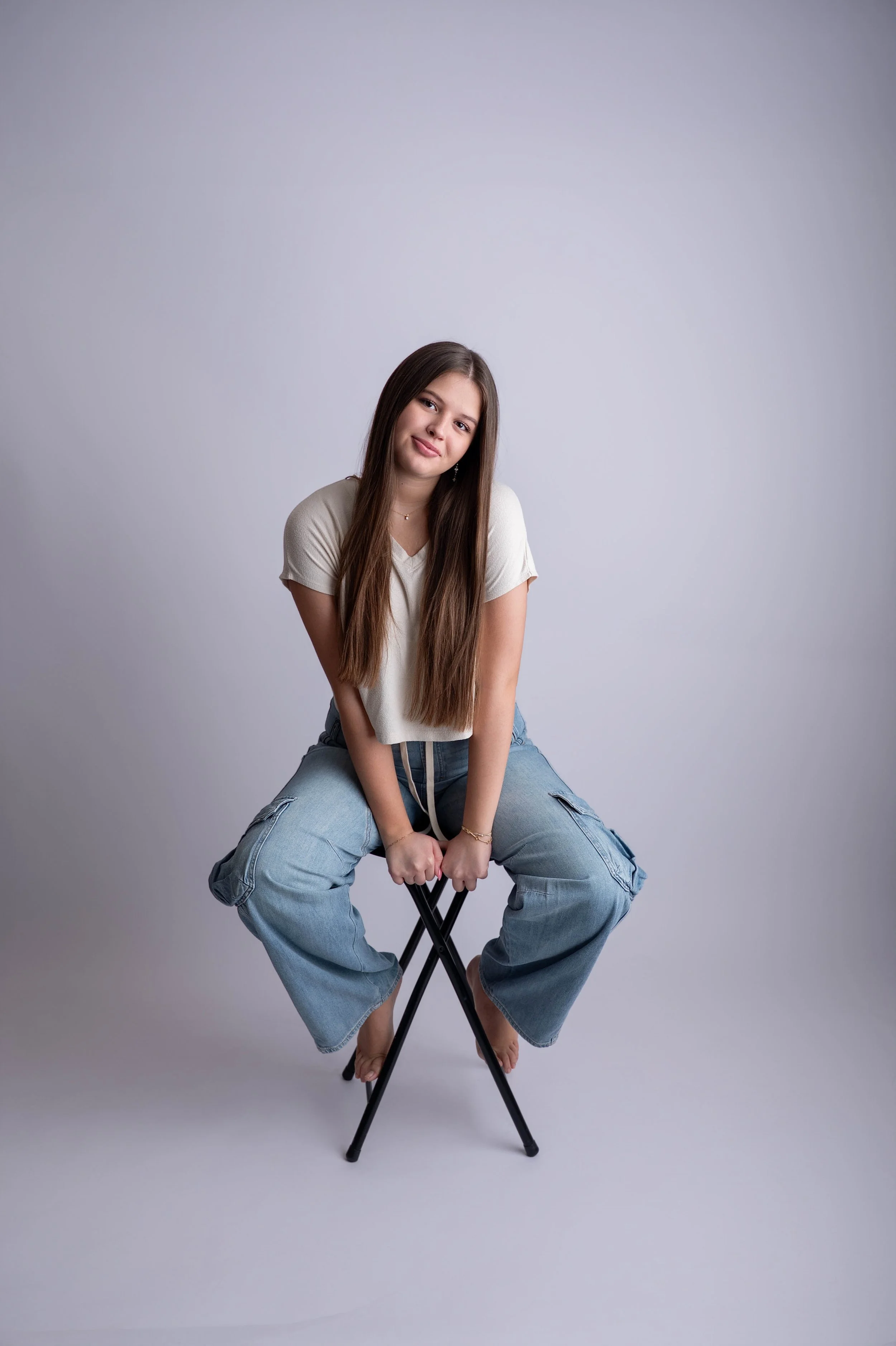 A young woman with long brown hair is sitting on a black folding chair, smiling softly, against a plain light gray background, wearing a white t-shirt and baggy blue cargo pants.