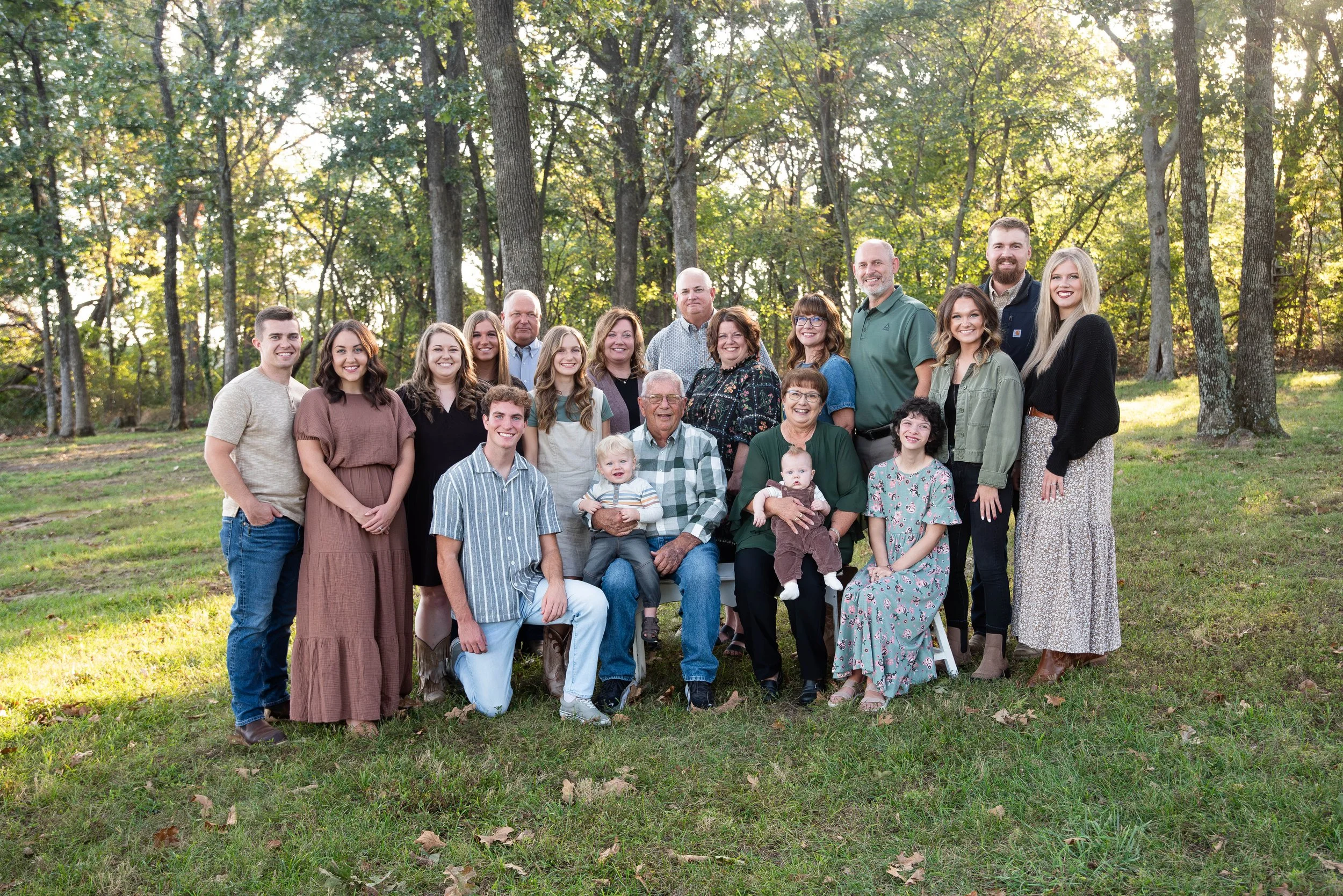 A large multigenerational family posing outdoors on a grassy area with trees in the background during daytime. The group includes older adults, middle-aged adults, young adults, teenagers, children, and babies, all smiling and dressed casually.